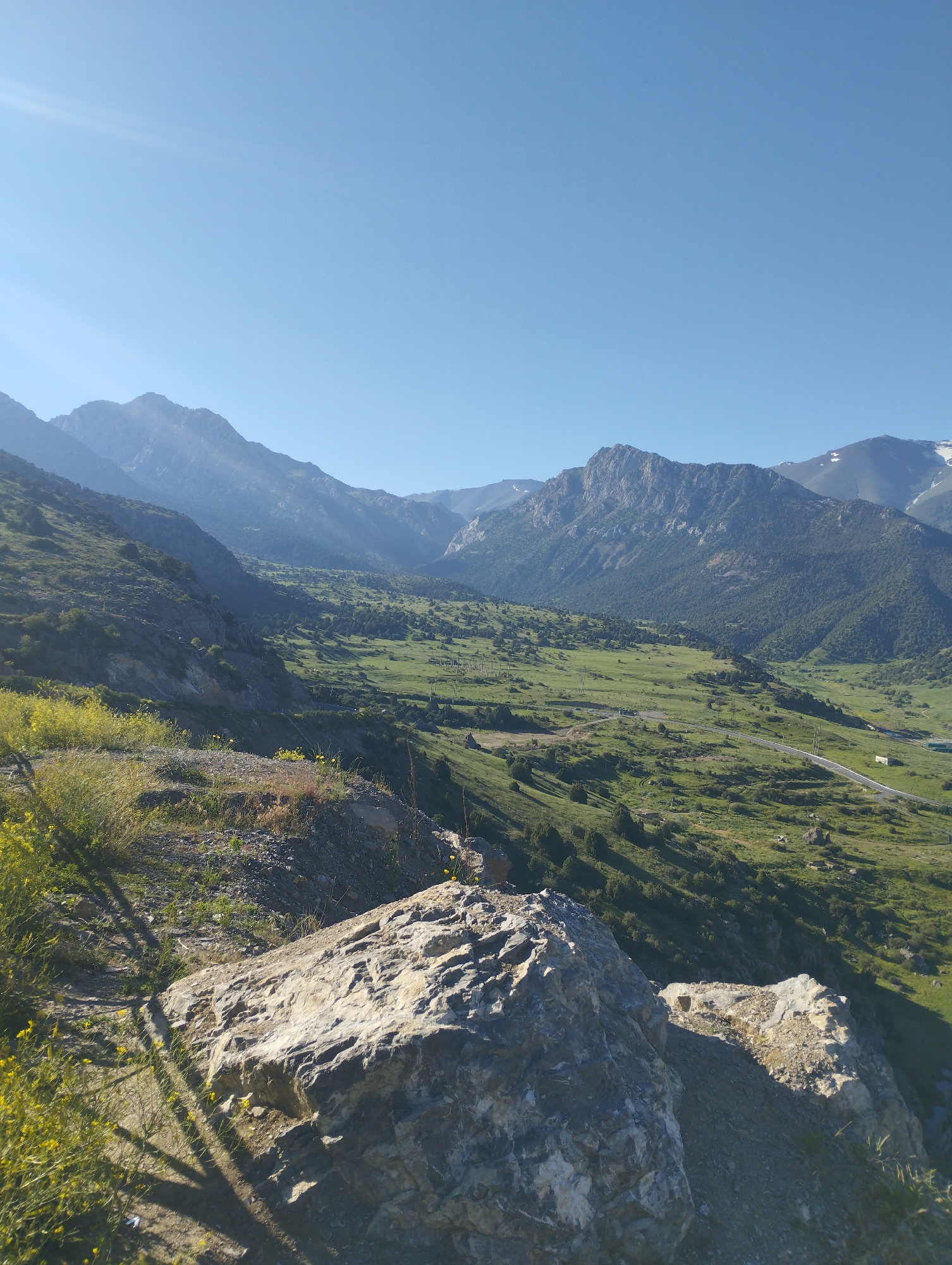 The image captures a scenic mountain valley landscape on a clear, sunny day. In the foreground, a large, textured rock is prominent, casting a shadow from a tree limb or structure. The foreground also features sparse green vegetation and small yellow wildflowers growing on a rocky incline.
The middle ground reveals a vast, verdant valley with rolling green hills dotted with dark green trees. A paved road snakes through the valley, with a few vehicles visible. The terrain is characterized by slopes and natural contours, suggesting a remote, natural environment.
The background is dominated by majestic, rugged mountains that recede into the distance. The peaks are sharply defined, with some appearing to have patches of snow or lighter rock at their summits. The sky above is a clear, bright blue, with a subtle sun flare visible in the upper left quadrant, indicating the direction of the sun.
The image is a landscape photograph, likely taken from a high vantage point, offering a panoramic view of the mountainous region. The location is identified as Dardar, Uzbekistan.