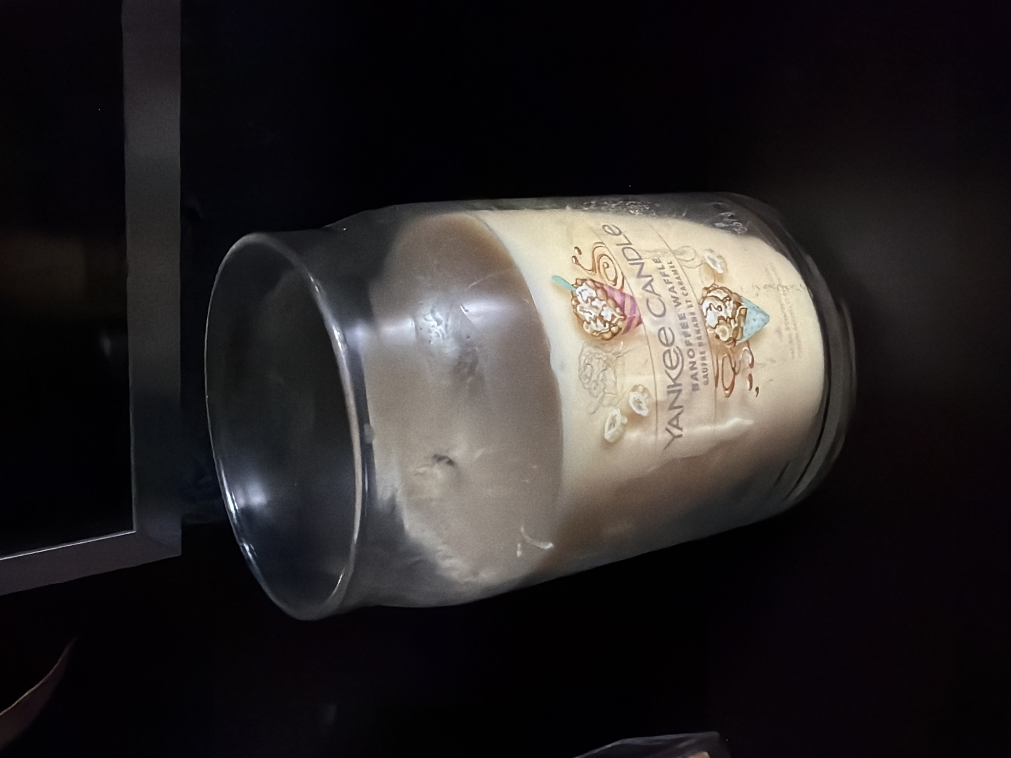 In an indoor, dark environment, a partially melted, light-colored candle sits in a clear glass jar. The candle is labeled with a "Yankee Candle" brand sticker, which also features graphics of waffles, ice cream, and caramel. The jar is tilted slightly to the right. The background is dark and indistinct, suggesting a shelf or furniture.