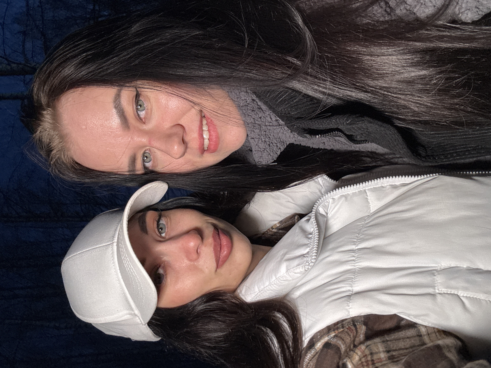 Two women are lying down. The woman on top has long, dark hair, fair skin, and is smiling. She is wearing a dark jacket and a gray sweater. The woman below is wearing a white baseball cap and a white puffer vest over a brown plaid shirt. Both women have light-colored eyes. The background is dark. The image was likely taken indoors. There is no indication that the image was taken in the city of Chaikovsky, Russia.