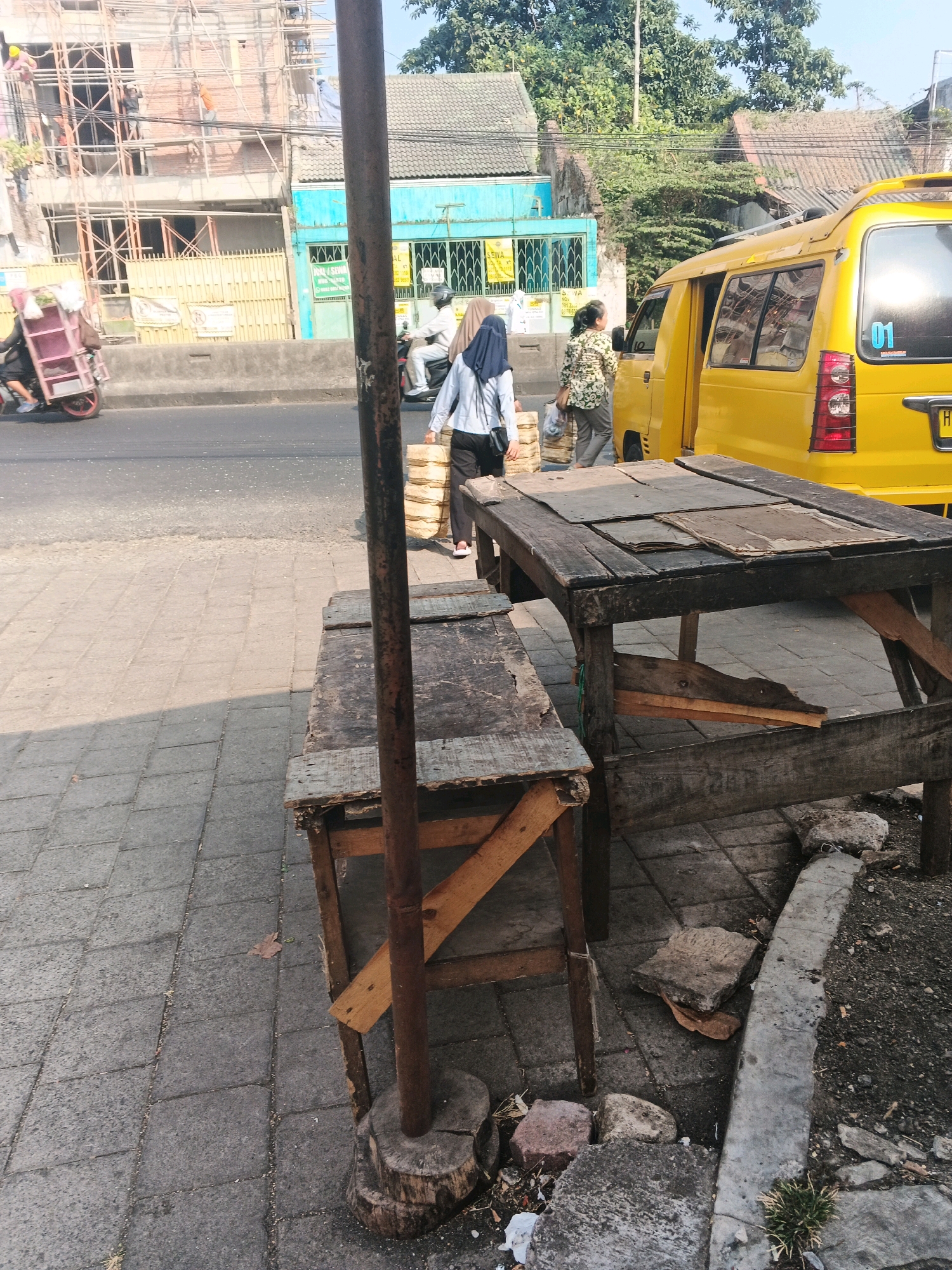 A view of a street scene in Ungaran, Indonesia, on a bright day. In the foreground, a rustic wooden table and bench sit on a tiled sidewalk. The table is covered with several flat, worn pieces of material. A metal pole, supporting some overhead wires, is embedded in a wooden base in front of the table and bench. Behind the furniture, a paved road is visible, with a yellow minivan parked on the right side. The minivan has the number "01" on its rear window. To the left of the minivan, two individuals are walking. One, wearing a light-colored hijab and a dark top, carries a stack of what appear to be packaged goods. The other, wearing a floral patterned shirt and dark pants, also carries a similar stack of items. Further back, a building under construction with scaffolding is visible, along with other buildings and trees. A motorcycle with a sidecar loaded with items is also seen on the road to the left.