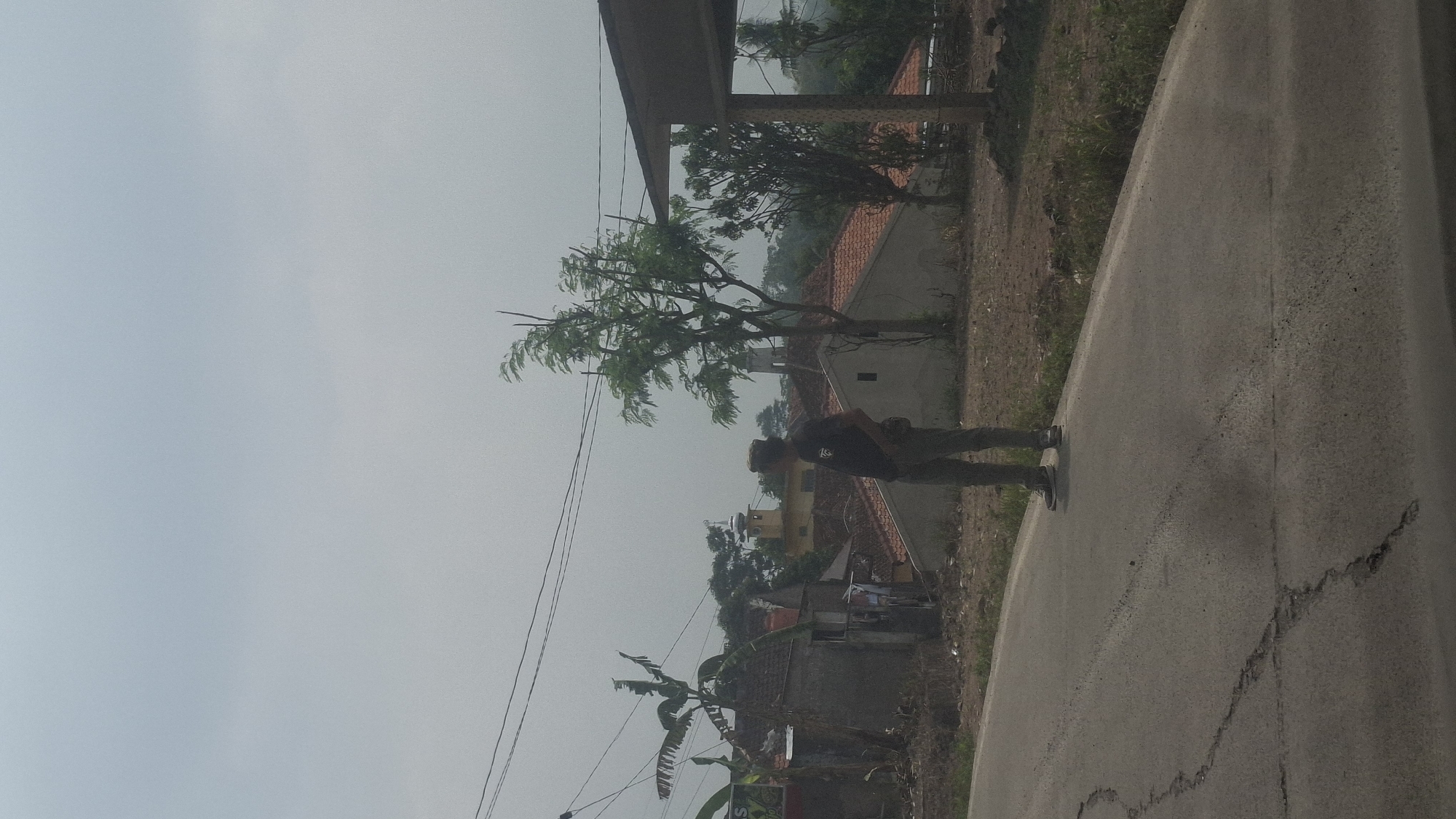 A full-body shot captures a person standing on the side of a road, looking towards the left of the frame. The individual is wearing a dark t-shirt and light-colored pants, with a hat and a dark bag slung across their chest. Their stance is relaxed, with hands in pockets. The road in the foreground is paved and cracked, with dry grass and dirt bordering it. In the background, a village scene unfolds. Houses with terracotta roofs are visible, along with a prominent yellow minaret of a mosque. A banana tree with large leaves stands to the left, and a leafy tree grows to the right of the person. Power lines are strung across the overcast sky. The scene appears to be a rural area in Sirnagalih, Indonesia, indicated by the presence of the mosque and the tropical vegetation.
