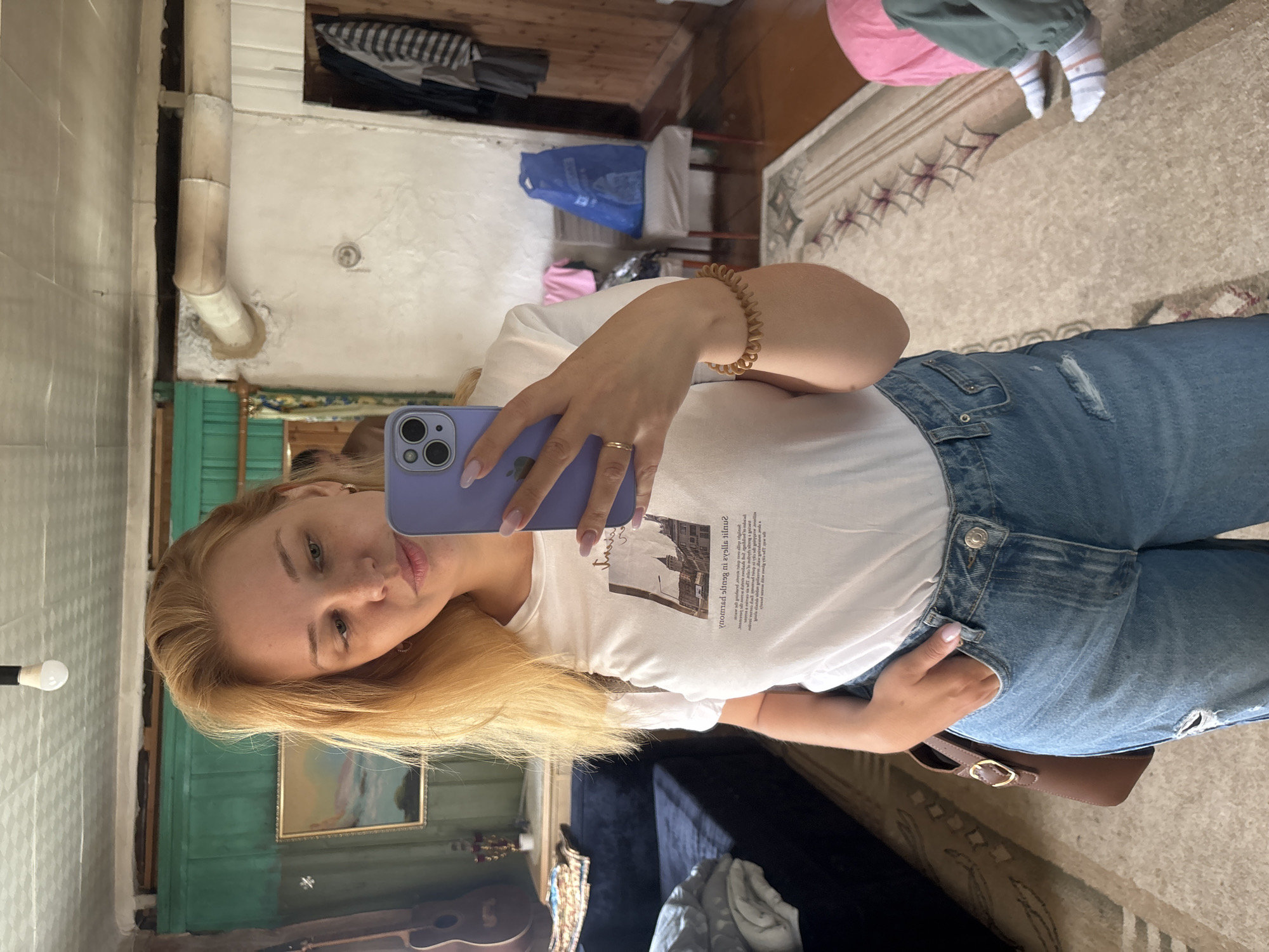 In Cherkasovo, Russia, a young woman with blonde hair stands in a dimly lit room, taking a selfie in a mirror. She is wearing a white t-shirt with a graphic print and light blue distressed denim jeans. Her left hand is tucked into her jeans pocket. She has a brown crossbody bag over her right shoulder. On her left wrist, she wears a spiral-shaped brown bracelet, and on her ring finger, a thin gold ring. The background reveals a cluttered room with a dark blue sofa on the left, an acoustic guitar leaning against the wall, and a painting on the green-painted wall. A large pipe runs along the upper right wall. Further into the room, a chair with a blue plastic bag on it is visible. The floor is covered by a patterned rug. In the reflection of the mirror, a person's legs are visible in the lower right corner, wearing striped socks and green pants, with a pink fabric nearby.