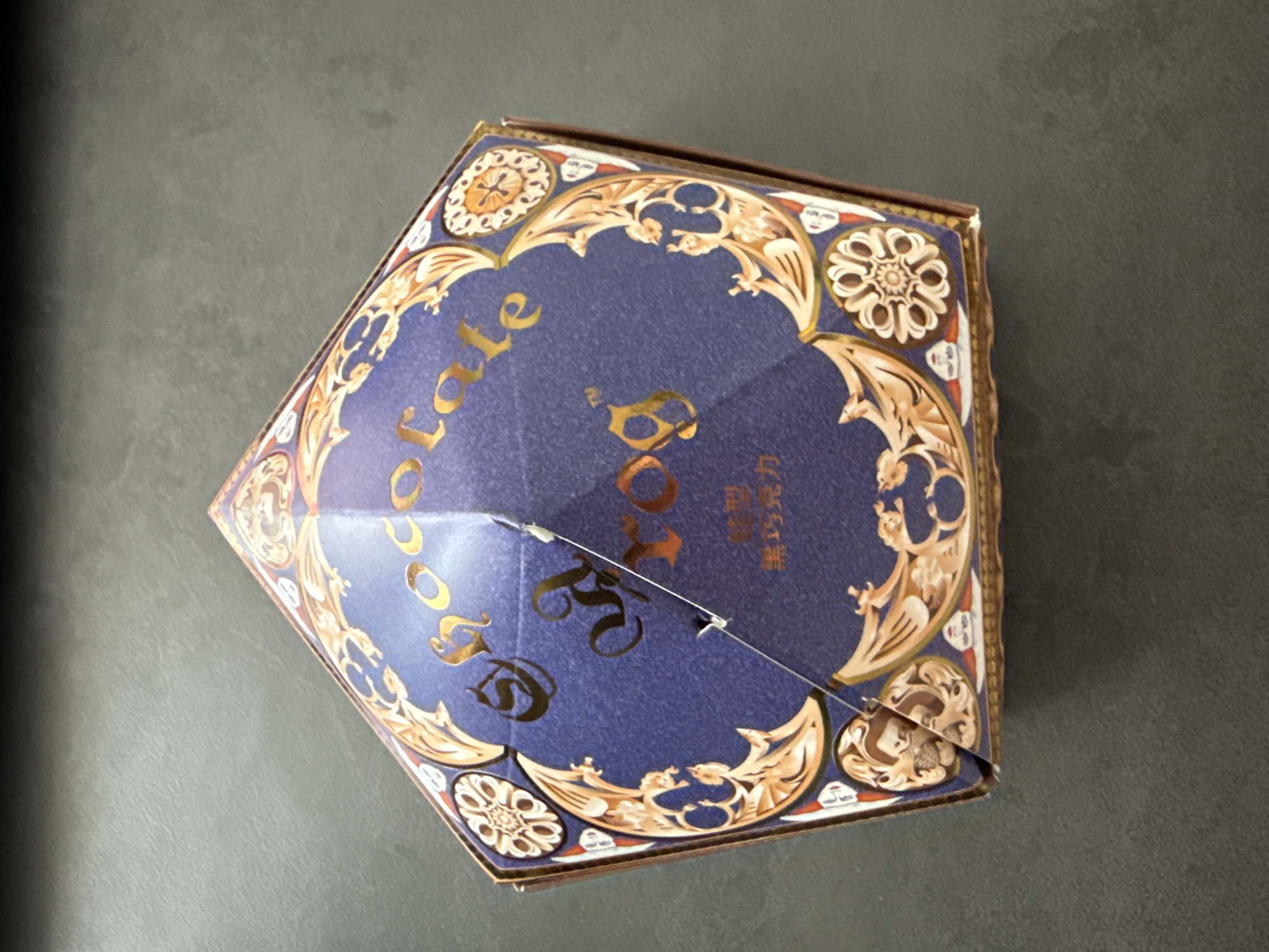 A top-down shot of a closed, pentagon-shaped chocolate box on a gray surface. The box is predominantly blue with gold lettering and an ornate gold design around the edges. The text on the box is "Chocolate Box". There is no indication of the location being Krasnoyarsk, Russia.