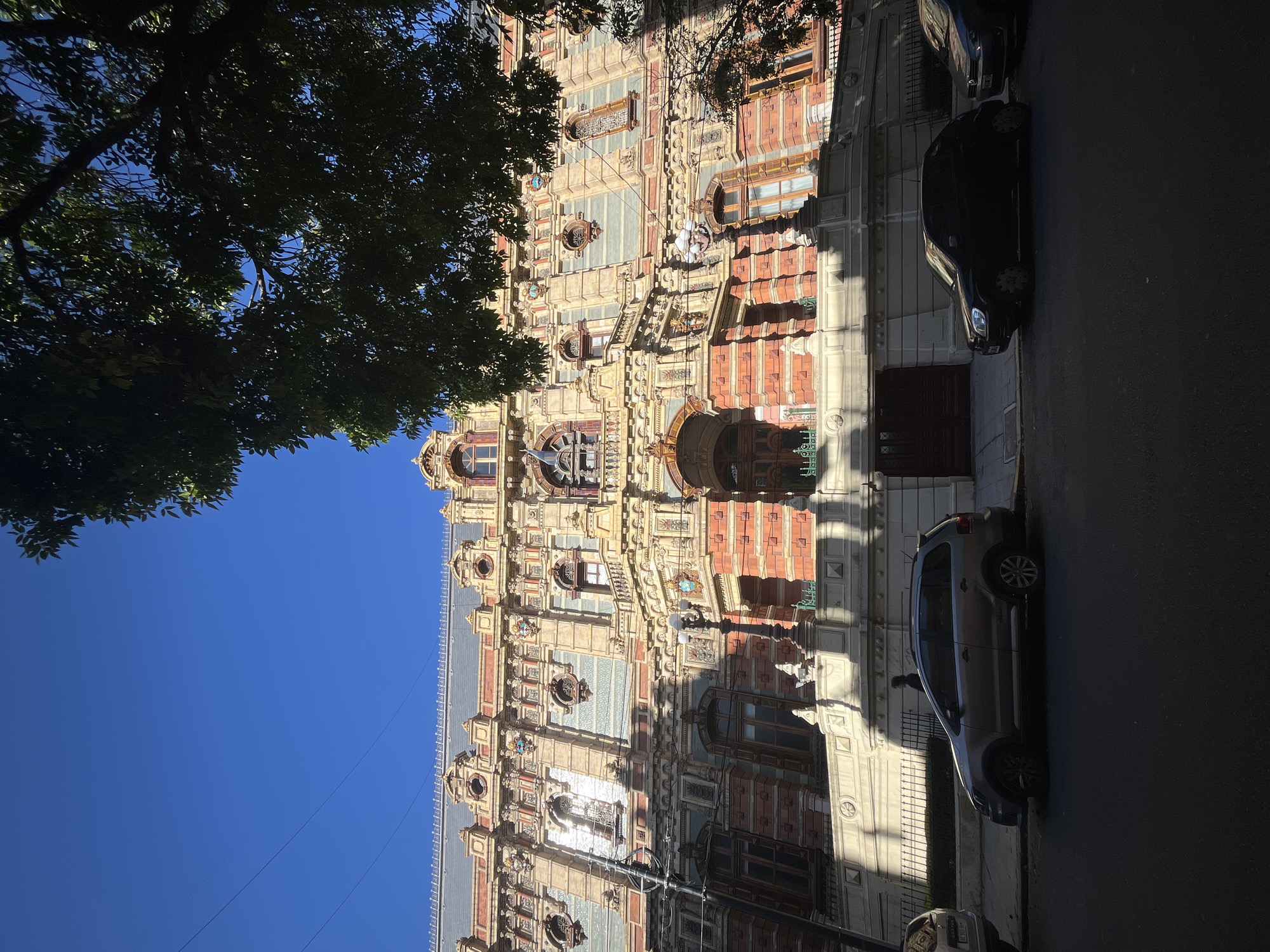 The image depicts the facade of the Palacio de Aguas Corrientes in Buenos Aires, Argentina. The building's intricate Beaux-Arts architecture is detailed with terracotta tiles and ornate decorations. The sky is clear and blue. A tree partially obscures the view on the left side. Two parked cars are visible along the street. The image is taken from a low angle, emphasizing the height of the building.
