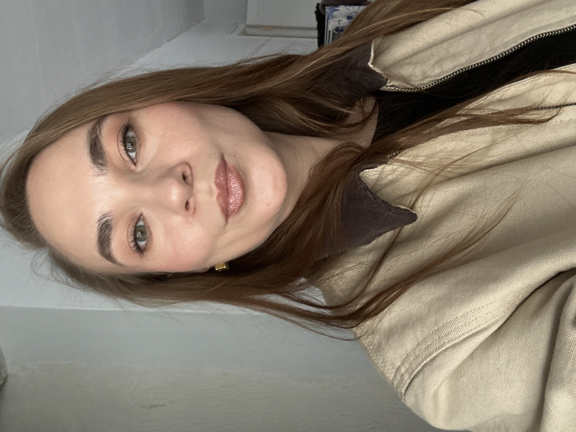 A woman with light skin, auburn hair, and green eyes is wearing a light tan jacket with a dark brown collar. She is looking at the camera with a slight smile. She is wearing a black top underneath the jacket and a small gold earring. The background is a plain white wall. The location cannot be determined from the image.
