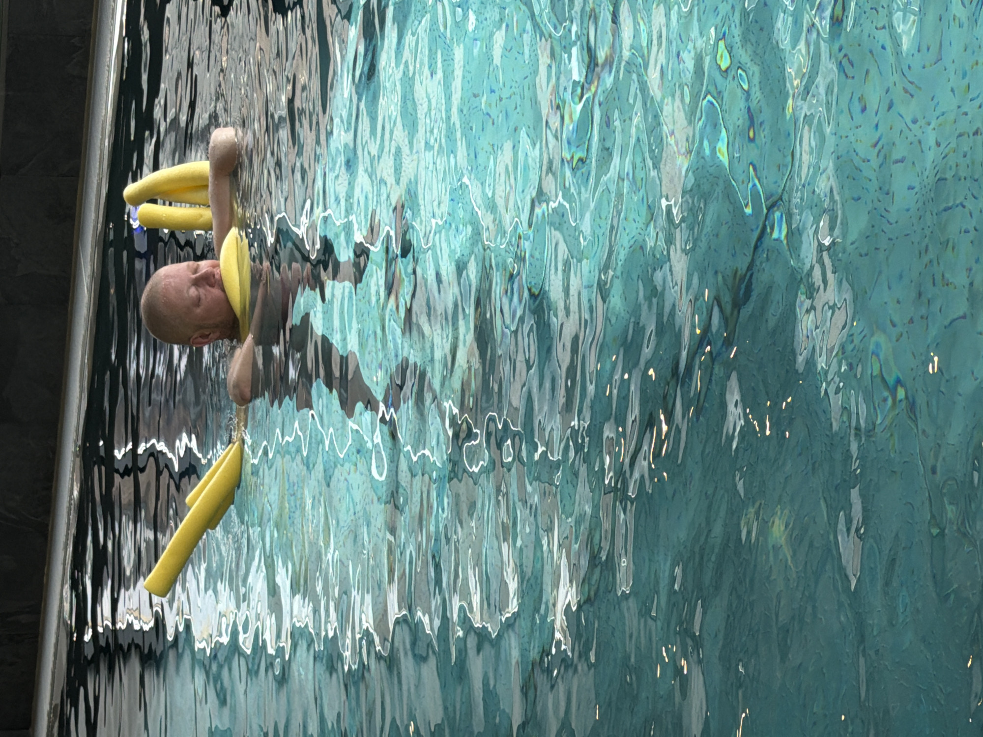 A man with a shaved head is in a pool, using two yellow foam noodles to float. He is holding one noodle behind his head and another in front of him. The water is turquoise and reflects light. The scene is located at a pool in the city of Chaikovsky, Russia. A portion of a wall is visible to the left.