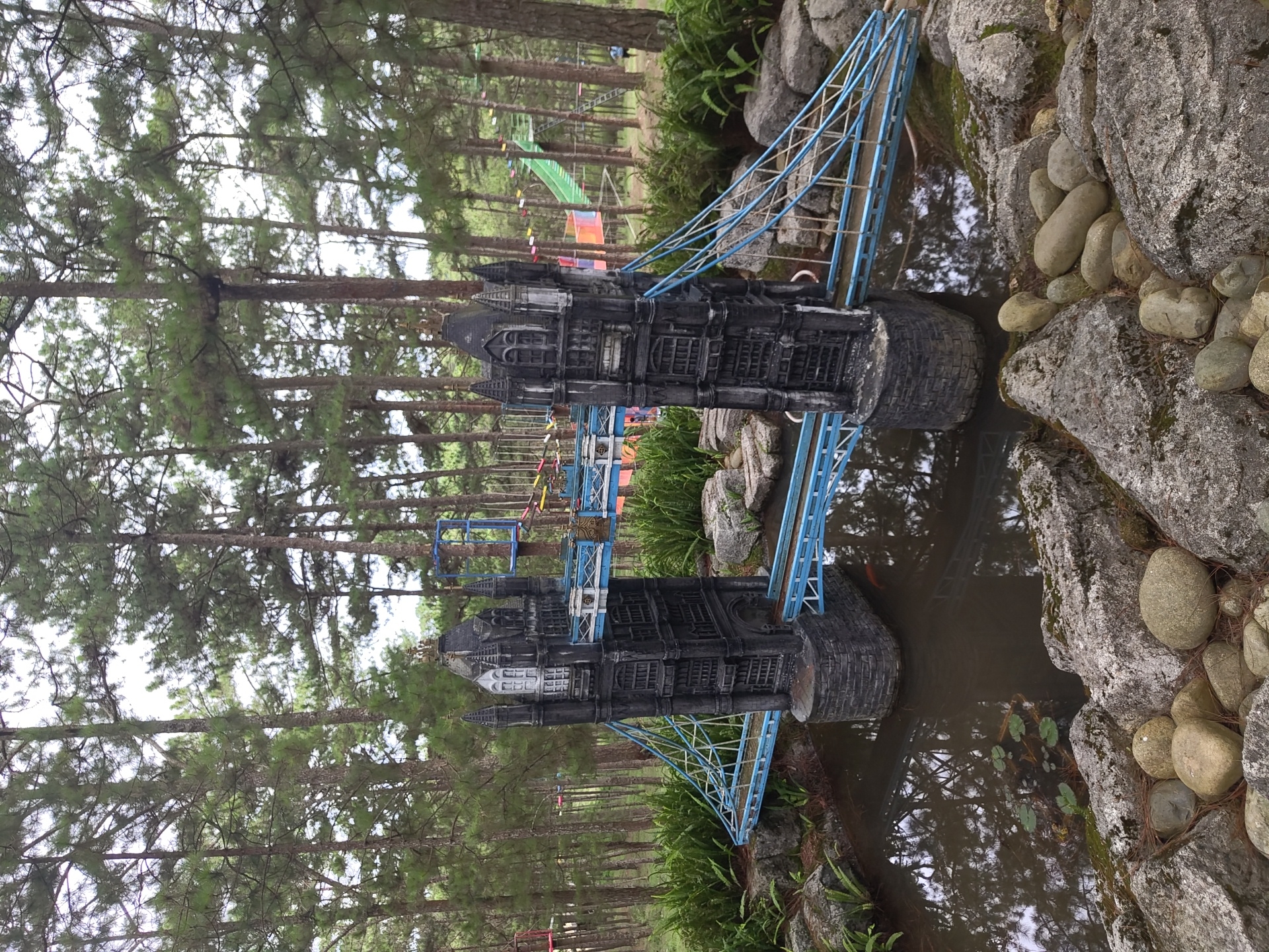 This image depicts a miniature replica of the Tower Bridge, a popular landmark of London, situated in the city of Lam Vien - Da Lat, Vietnam. The model bridge, constructed with dark grey stone-like material and a bright blue suspension system, spans a small body of water. The water reflects the surrounding pine trees and the sky, with a few lily pads visible on its surface. In the water, a small orange fish can be seen. The bridge is flanked by lush green ferns and moss-covered rocks in the foreground, contributing to a naturalistic setting. In the background, tall pine trees dominate the landscape, suggesting a forested or park-like environment. A colorful rope course or adventure park structure is also visible in the distance, with a bright green slide and various ropes and platforms. The overall scene suggests a themed attraction or garden designed to replicate famous landmarks within a natural Vietnamese setting. No people are clearly identifiable in the image.