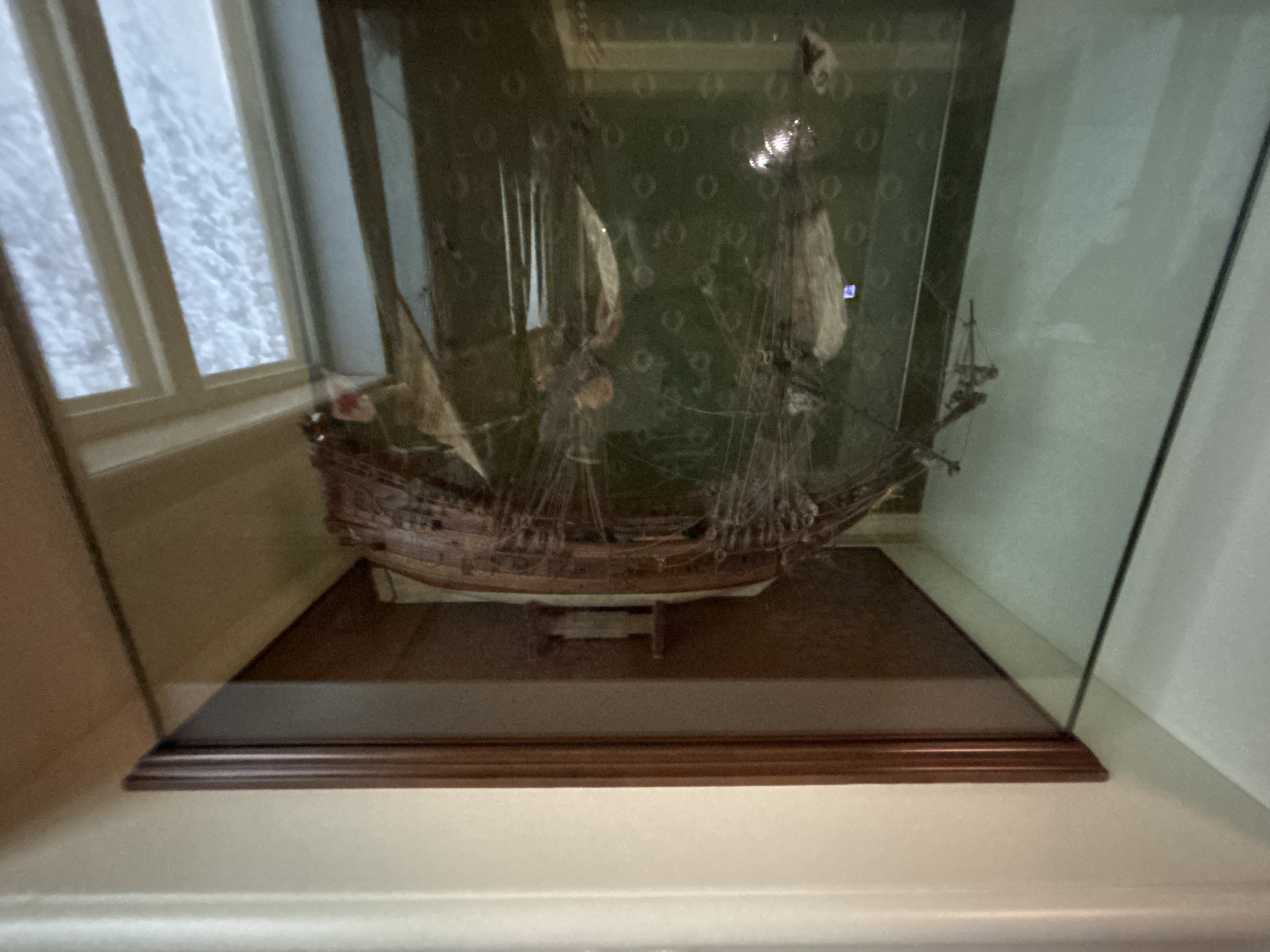 The image captures a detailed model of a multi-masted sailing ship, possibly a galleon, displayed within a highly reflective glass case. The model ship is constructed from dark wood, featuring intricate rigging and light-colored, textured sails, and is mounted on a small stand. Behind the ship, within the display, a dark green wall with a repeating pattern of light circular motifs is visible. Significant glare and reflections on the glass obscure parts of the model. A prominent reflection on the left side of the case shows a multi-paned window looking out onto a bright, textured, whitish-grey scene, possibly a snow-covered landscape or cloudy sky. A small, bright blue rectangular reflection is visible on one of the ship's sails. The display case has a dark brown wooden base with a subtle molding, resting on a lighter, off-white wall or pedestal in the foreground. No individuals are present in the static scene, which is located in an indoor exhibition or museum setting in the city of Yekaterinburg, Russia.