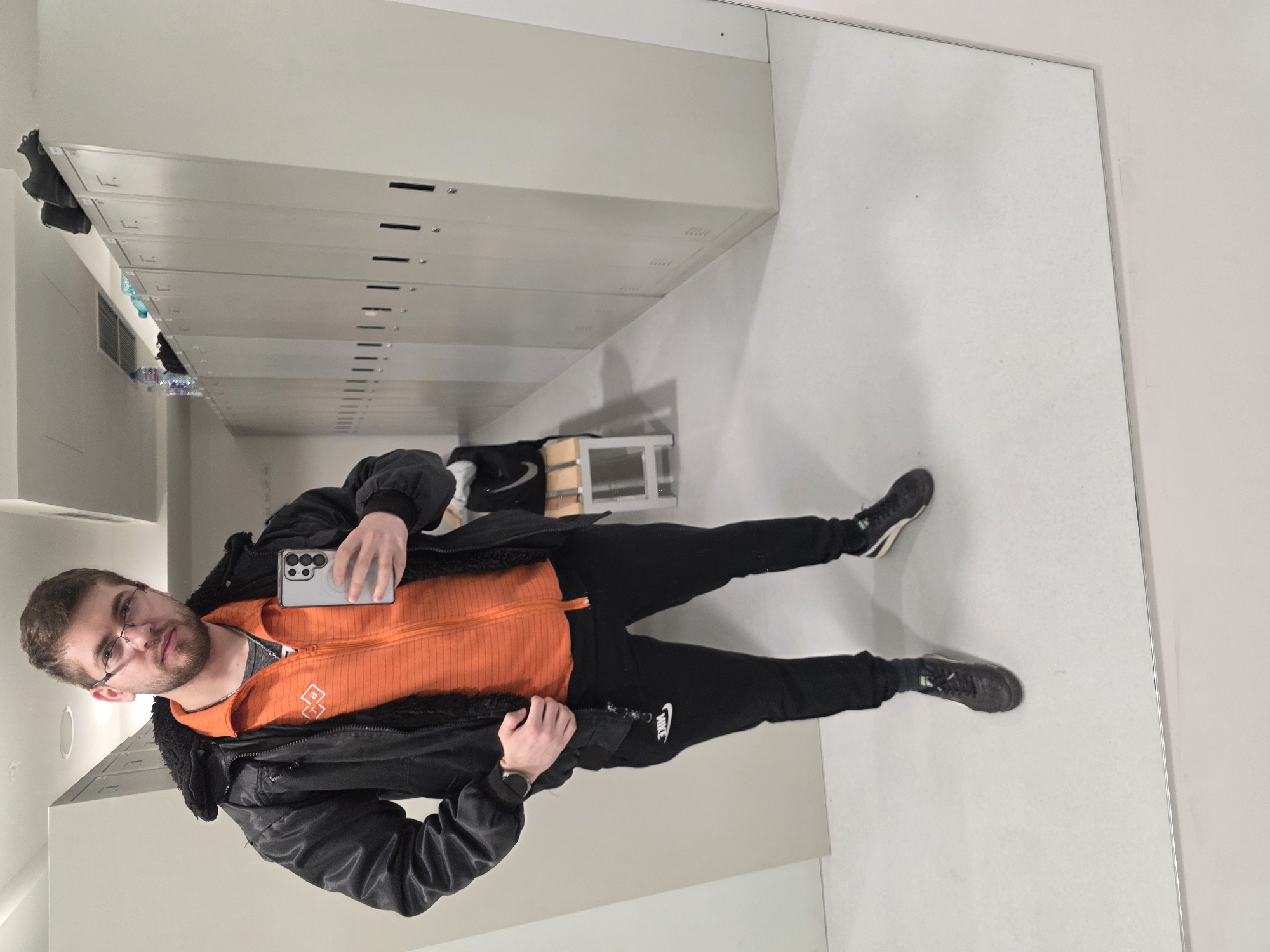 A man is seen taking a selfie in a mirror. The man is wearing glasses, a black jacket, an orange sweater, and black Nike sweatpants. He is holding a smartphone in his left hand. The environment appears to be a locker room. Silver lockers line the wall. The location is not identifiable as Plovdiv, Bulgaria based on the image alone.