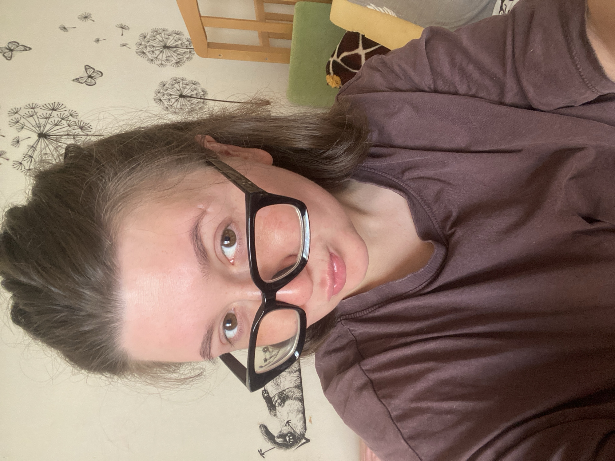 A young woman wearing glasses and a dark purple t-shirt is positioned in front of a patterned wall featuring dandelion and butterfly designs. The image is taken at an oblique angle, with the subject’s head tilted slightly. In the background, there is a partially visible wooden chair frame and a green cushion with a brown patterned object on it. A drawing of a squirrel is seen on the wall behind her glasses. Based on the user's prompt, this image was taken in Chaikovsky, Russia.