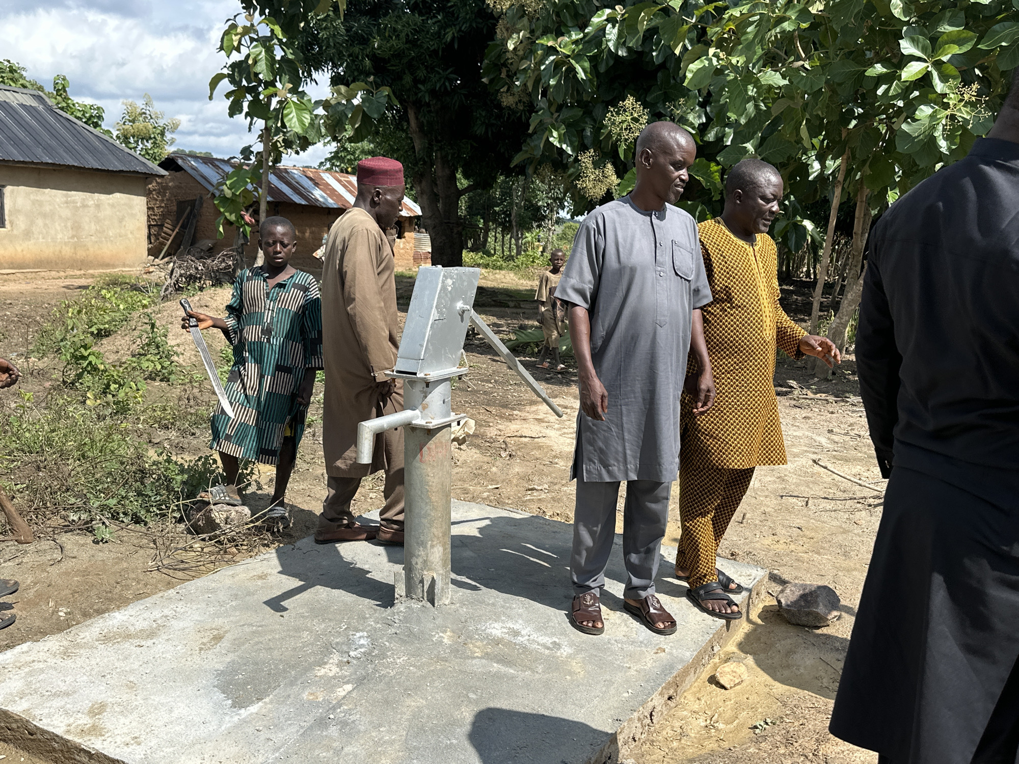 The image captures a scene in Afogo, Nigeria, focused on a newly installed water pump. Four individuals are present, with three men in the foreground gathered around the pump. The man on the far left, wearing a gray tunic and trousers, is facing away from the camera. Next to him, a man in a patterned yellow and black robe points towards the pump. To his left, a man in a brown tunic and red cap stands looking towards the pump. A young boy in a patterned green and black garment stands to the left of the pump, holding what appears to be a metal tool. The environment is rural, with a dirt ground, vegetation, and buildings with corrugated metal roofs visible in the background. The sky is cloudy. The water pump is constructed of metal and is mounted on a concrete base. The overall impression is one of a community project or an inspection of a new facility.