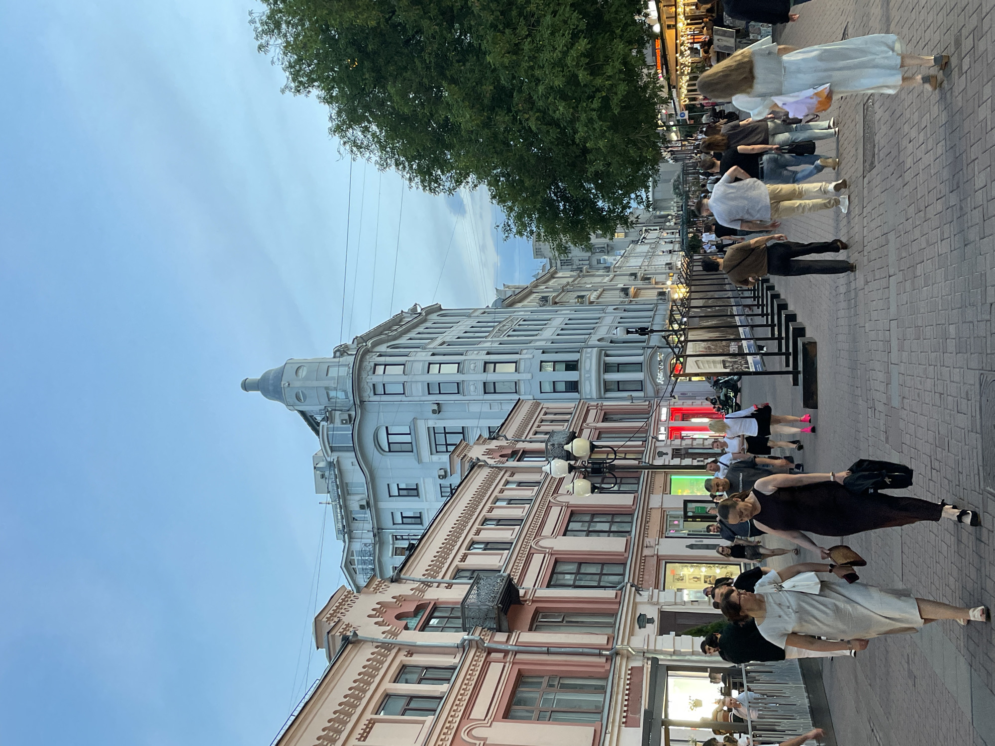 The image depicts a street scene in Moscow, Russia. The buildings lining the street are a mix of architectural styles, including a prominent grey building with a turret. People are walking along the paved sidewalk. Some are dressed in casual attire, while others are more formally dressed. Street lamps line the sidewalk. A tree is visible in the upper portion of the image. The sky is overcast.