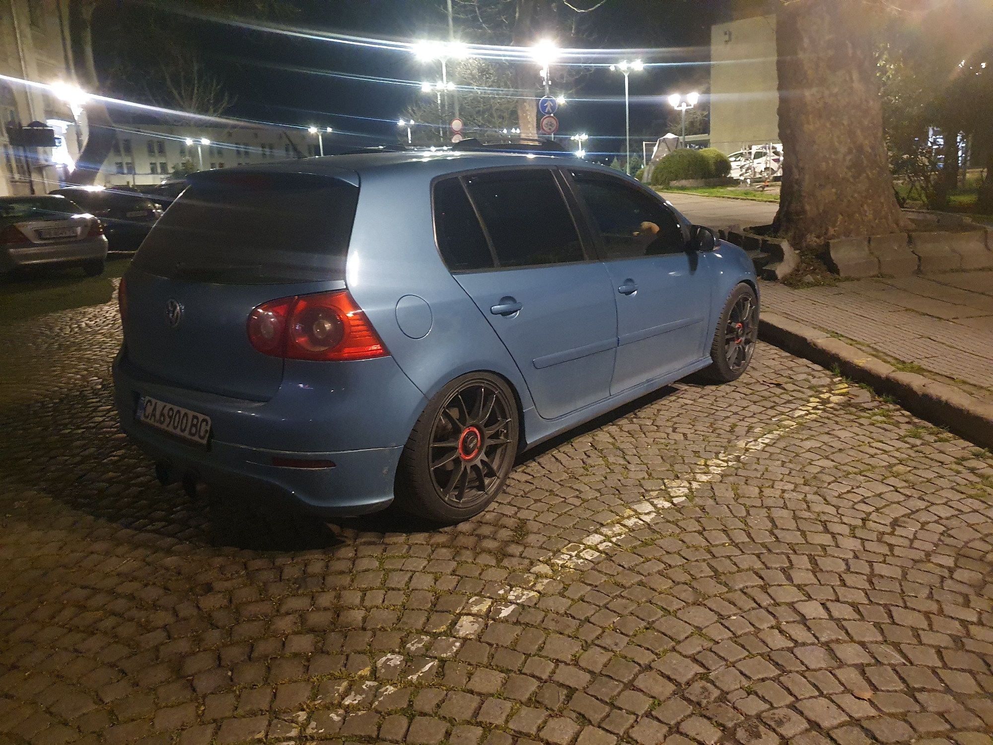 A blue Volkswagen Golf is parked on a cobblestone street in Plovdiv, Bulgaria. The car is facing right, with its rear visible. The license plate reads "CA 6900 BG." Behind the car are other parked cars and a building. Street lights illuminate the scene. To the right of the car is a large tree and a sidewalk.
