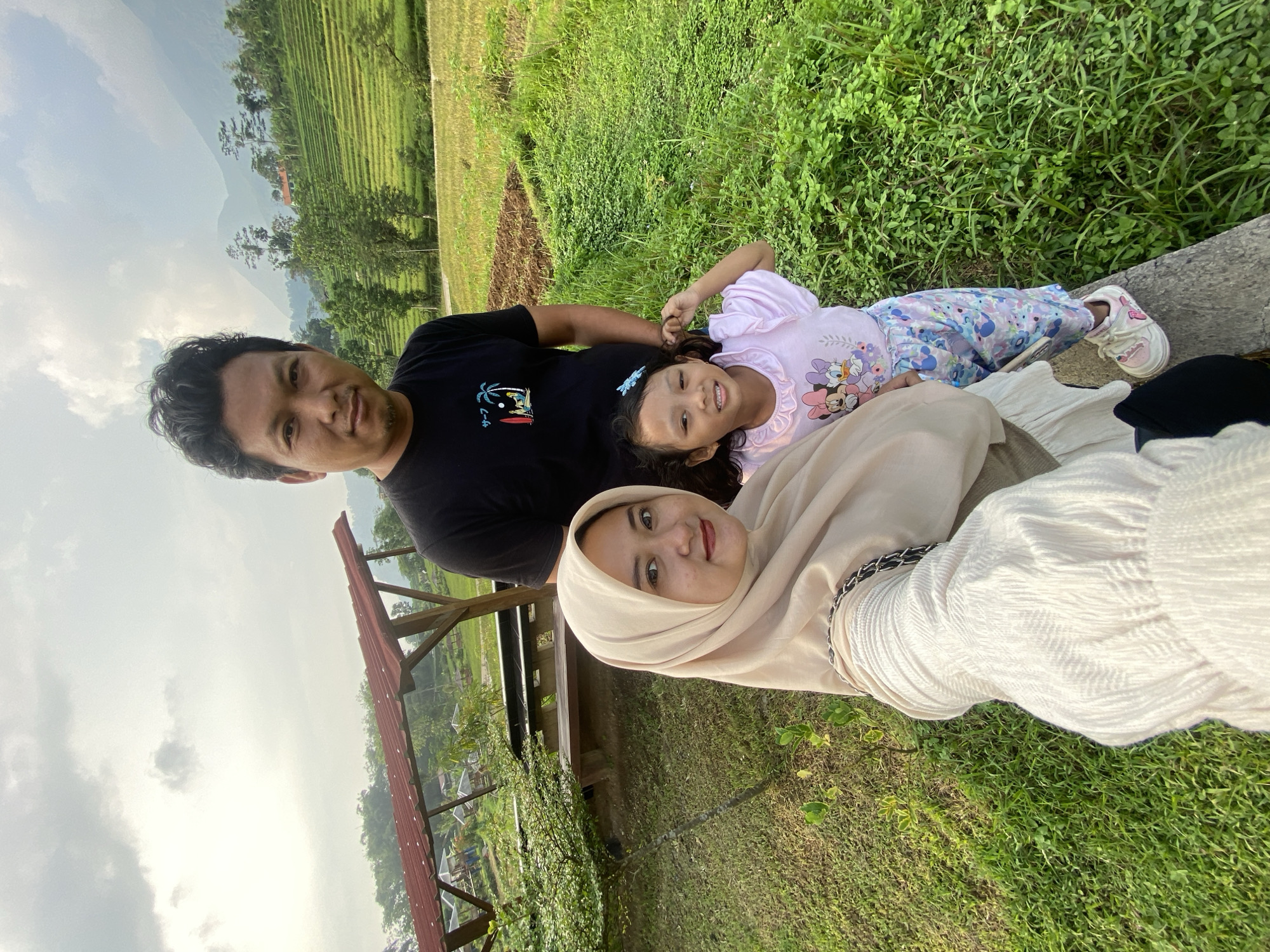 Three individuals—a man, a woman wearing a hijab, and a young girl—are situated outdoors against a backdrop of lush greenery and terraces, potentially rice terraces, near Mount Penanggungan in Indonesia. The man is wearing a black t-shirt; the woman is wearing a light-colored hijab and a white patterned top; and the child is lying on the man's stomach, wearing a pink top and patterned pants. The ground is covered with grass and vegetation. A wooden structure is visible on the left, seemingly part of the landscape. The photo seems to be taken from a low angle, focusing on the subjects against the panoramic background.