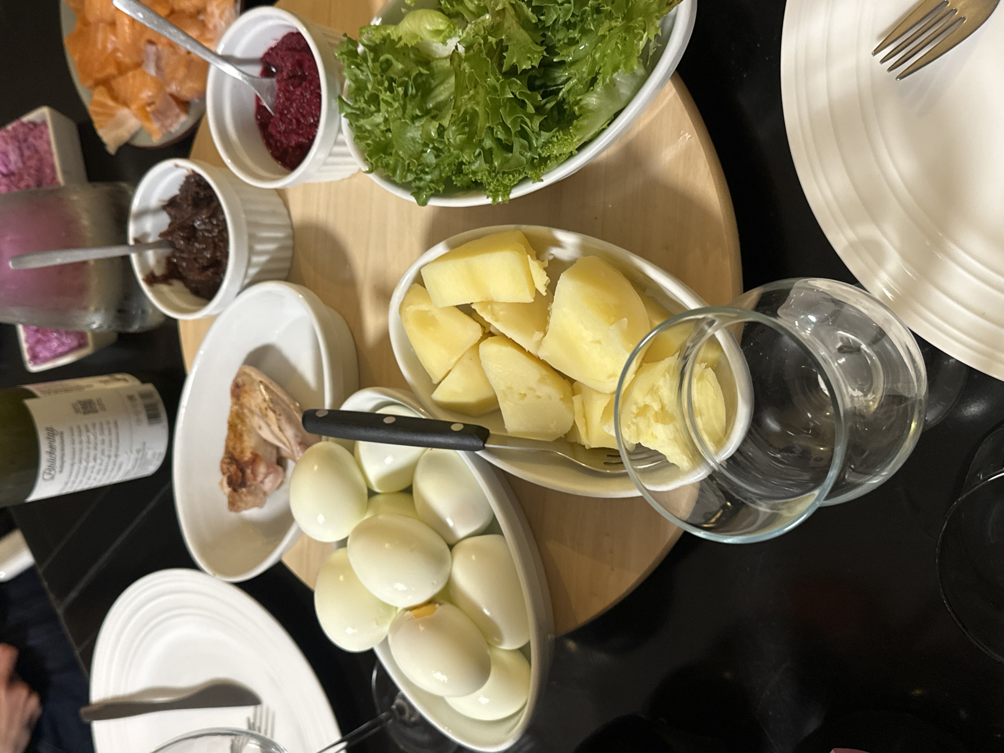 The image shows a table setting with various dishes of food, likely in a home in Ramat Gan, Israel. Dishes include boiled potatoes, hard-boiled eggs, salad greens, herring, chopped liver, gefilte fish, beets, and salmon. There are plates, forks, knives, wine glasses, and a bottle of white wine present. The scene suggests a meal is about to be, or is being, served. A person's feet can be partially seen in the bottom left corner. The table has a black surface and a wooden cutting board is present.