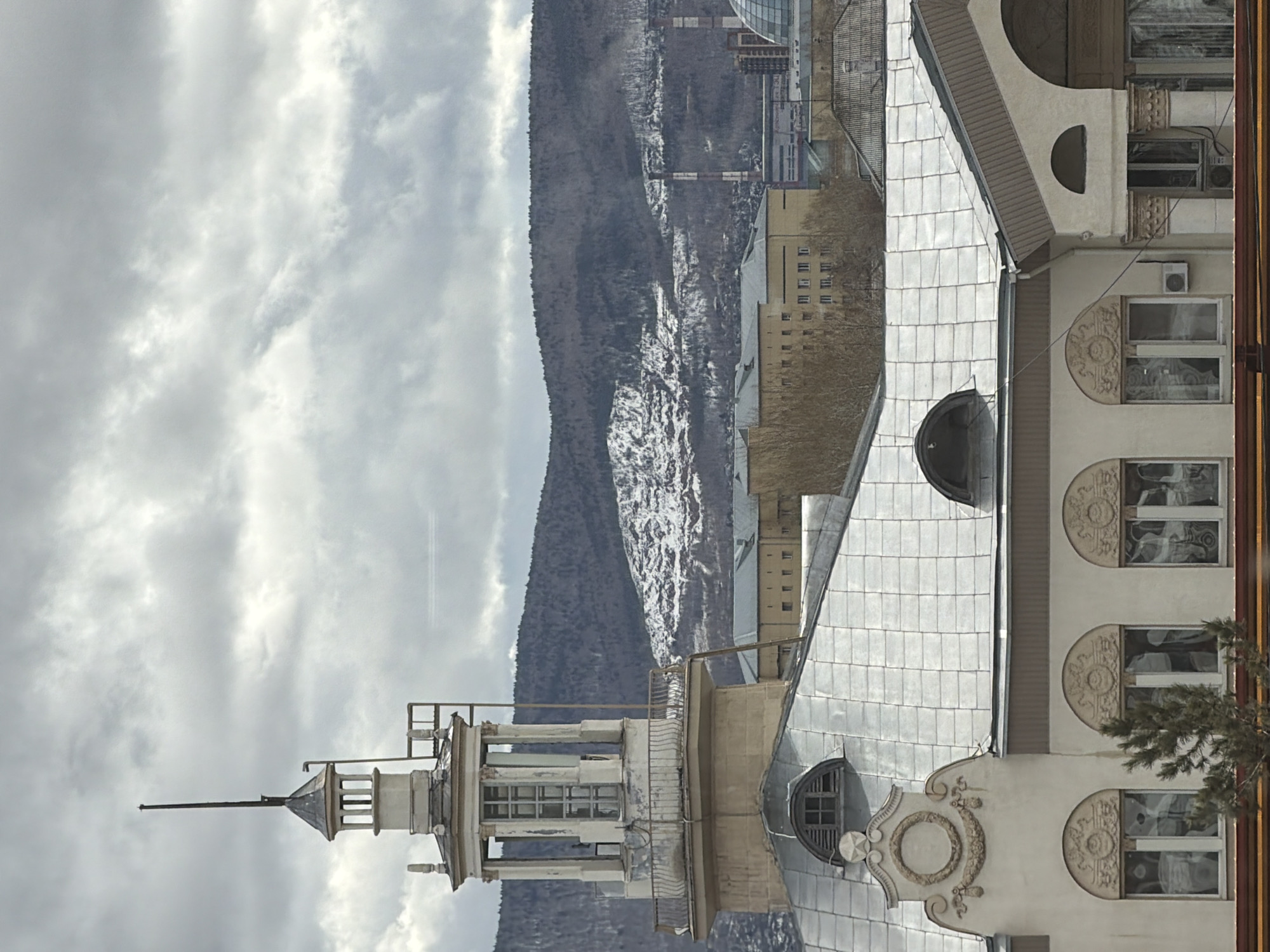 The image depicts a view in Krasnoyarsk, Russia, featuring a building with a distinctive turret or spire-like structure, suggesting it may be of historical or architectural interest. A mountain range is visible in the background. The sky is overcast with heavy clouds. The architectural style of the building is ornate, featuring rounded windows and decorative molding.
