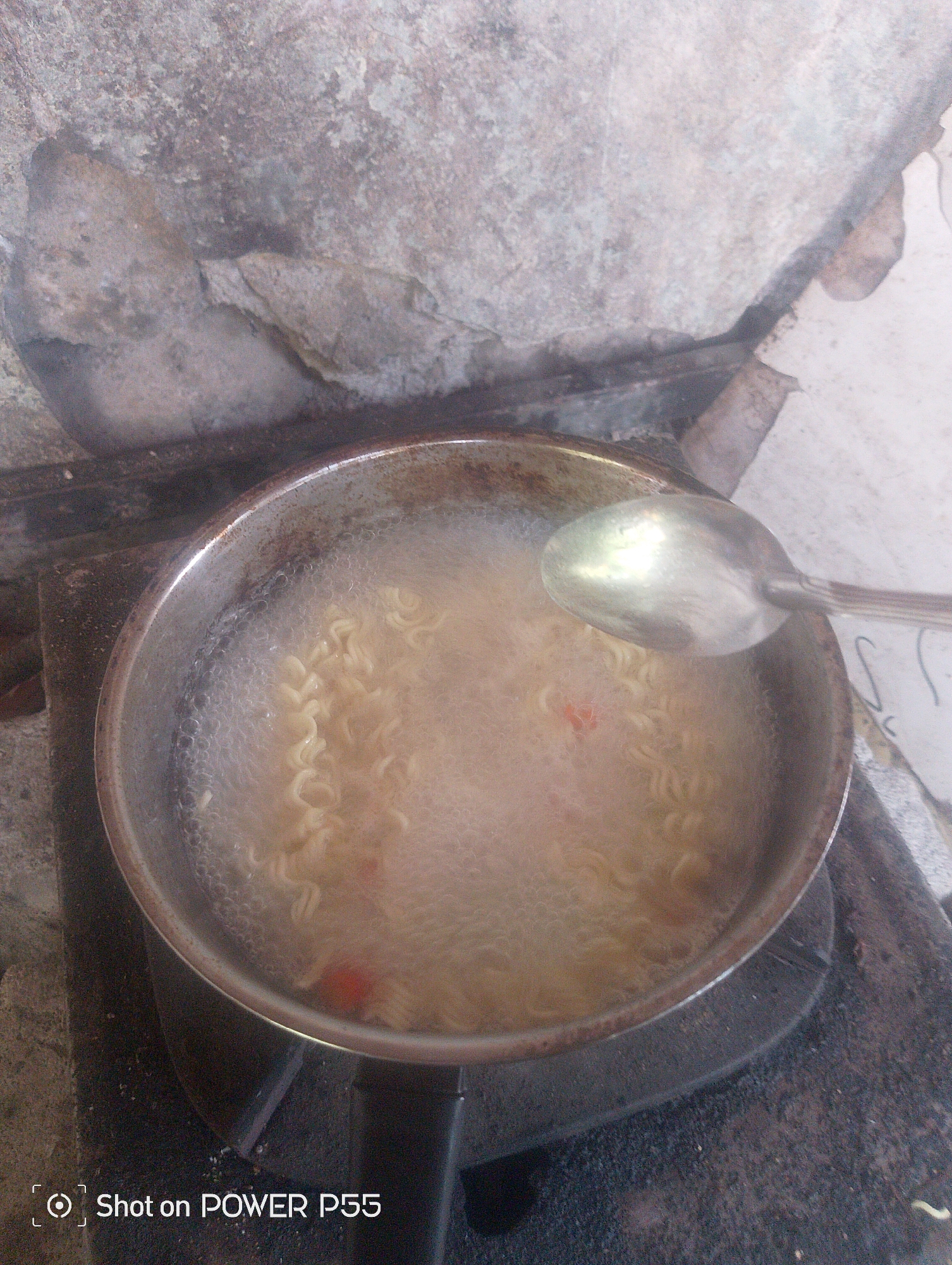 A metal pot filled with bubbling water and instant noodles is placed on a rustic outdoor stove, with a spoon resting on the rim of the pot. In the background, a textured, weathered wall suggests an outdoor setting in Lebaksiu, Indonesia. The image is captured from a top-down perspective, with the lower portion of the frame displaying a watermark indicating it was taken with a POWER P55 phone.