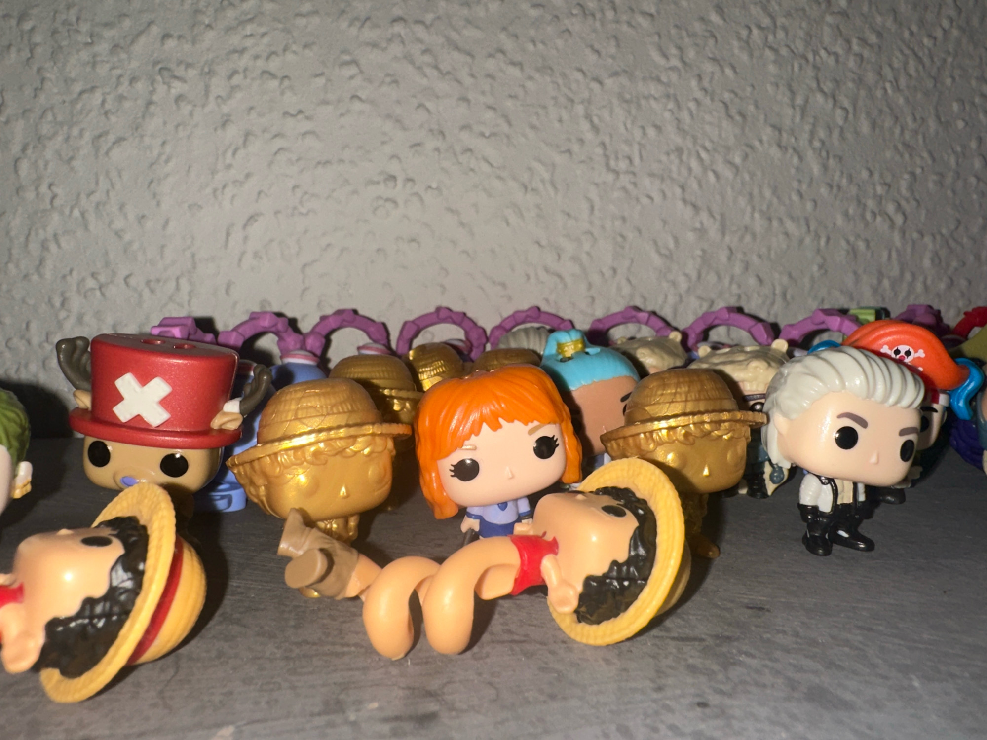 The image shows a collection of Funko Pop figures arranged on a shelf. The figures are mostly characters from the anime and manga series One Piece. In the foreground, a figure with a stretched, flexible neck is contorted, with its head resting inside a straw hat. Other figures visible include characters with red hats, gold hats, and white hair. In the background, there are more figures with purple accessories. The figures are placed on a grey surface, and the wall behind them has a textured, light-colored finish. This image does not depict any specific location in the city of Günzburg, Germany.