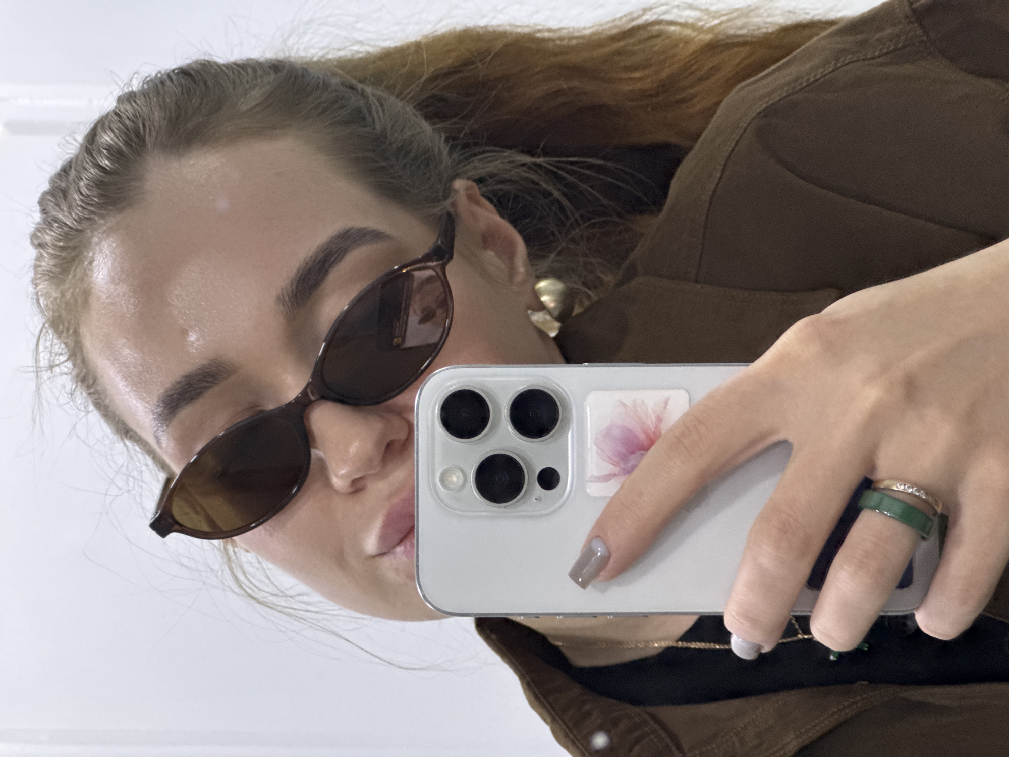 A person with brown hair is holding a white smartphone to take a selfie. They are wearing brown sunglasses and a brown jacket. The phone case has a floral design. They have rings on their finger, one is gold with embedded stones, and the other is green. The location is not specified.