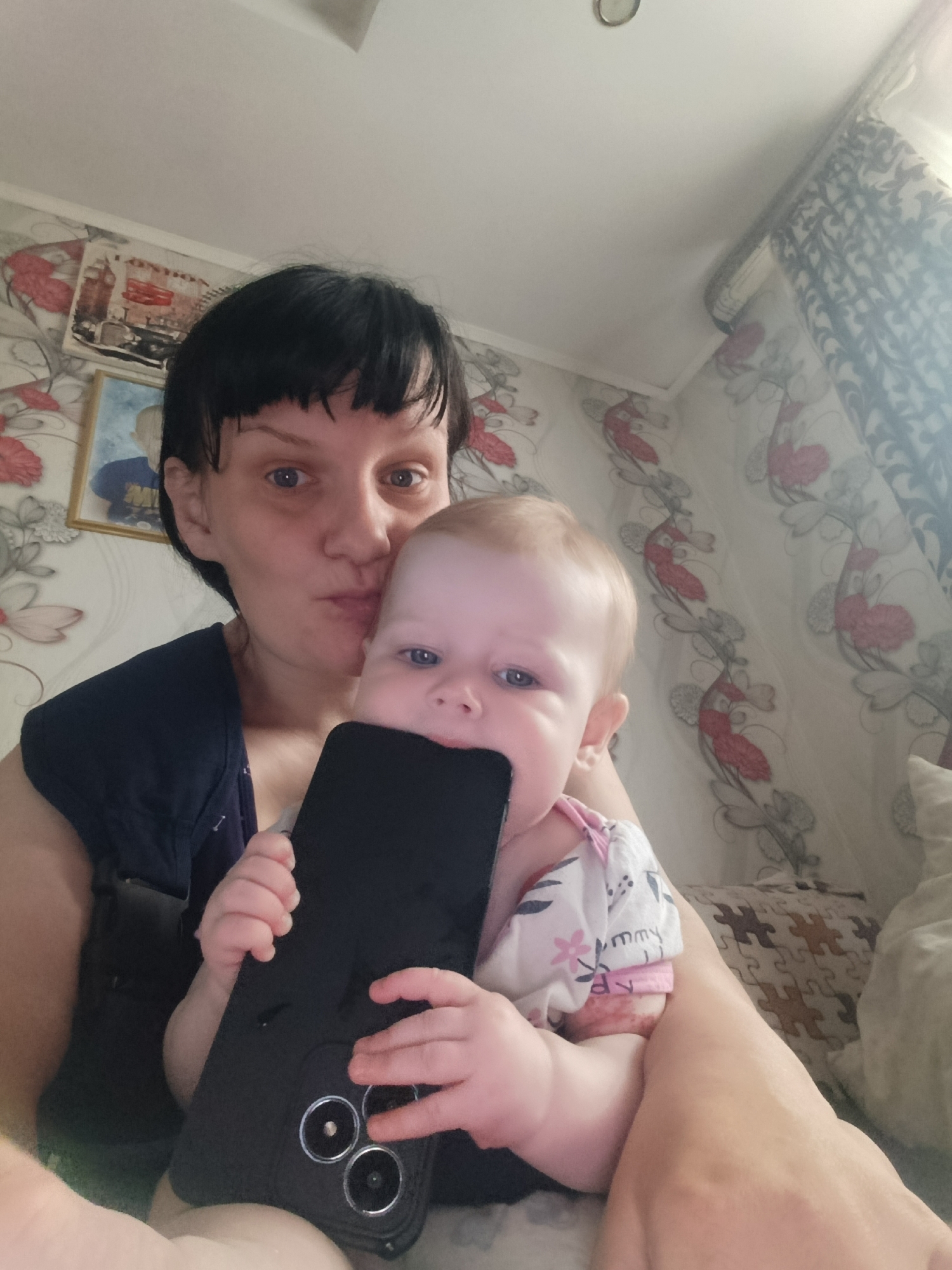 A woman is holding a baby who is biting on a cell phone. The woman has short, dark hair and is wearing a navy blue shirt. She is looking at the camera with pursed lips. The baby has fair skin and light hair, and is wearing a white and pink patterned outfit. The cell phone is black and has three circular camera lenses visible on the back. They are in a room with floral wallpaper, a framed picture of London, England, and patterned curtains. The location is Unknown, Unknown.