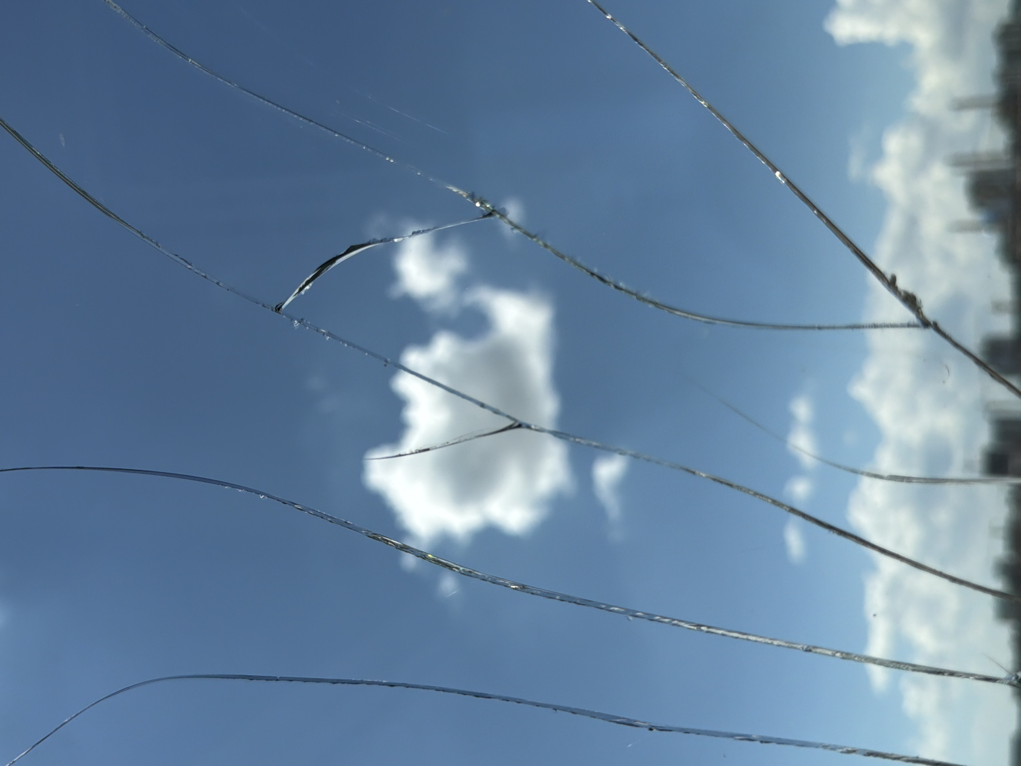 The image shows a fractured glass surface against a bright blue sky with scattered white clouds. The cracks in the glass are prominent, creating a network of lines across the frame. A single, puffy white cloud is visible in the center, while a distant, blurred cityscape with buildings and structures is visible at the bottom of the frame. The light appears to be natural daylight, suggesting an outdoor scene. The image does not provide any specific identifiable landmarks or context to pinpoint its location within the city of Astana, Kazakhstan.