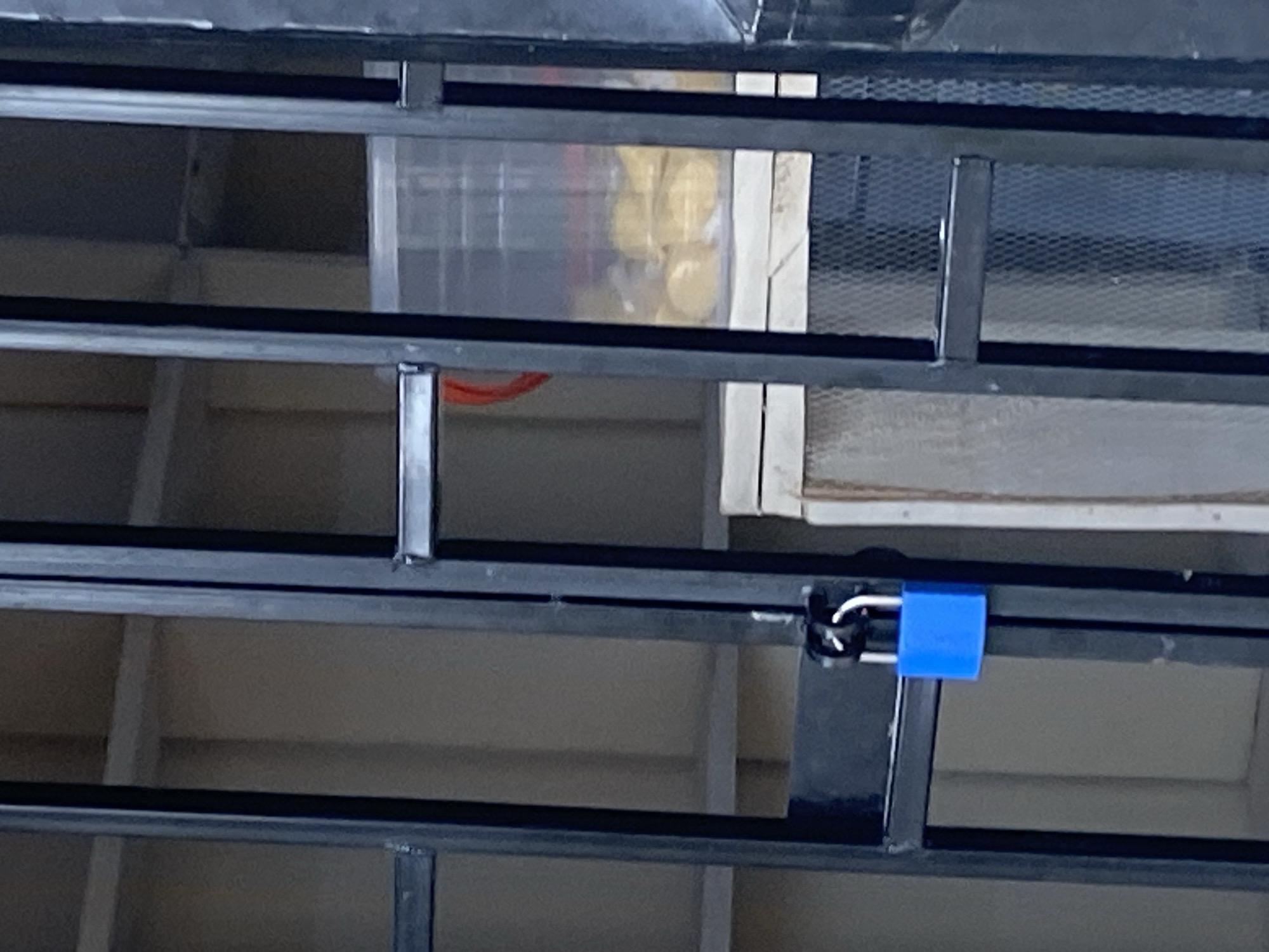 The image shows a close-up view of a black metal gate or fence with a bright blue padlock securing it. The gate appears to be made of vertical bars and a horizontal crossbar. Behind the gate, shelves or partitions are visible, along with a clear plastic container holding yellow, round objects, possibly food items. The background suggests an indoor or covered outdoor area, possibly a shop or storage space. There are no people or animals present in the image. The scene is static and well-lit, with no indications of any activity. The location is identified as Jauro Yinu, Nigeria, but this context does not offer specific visual cues within the image itself.