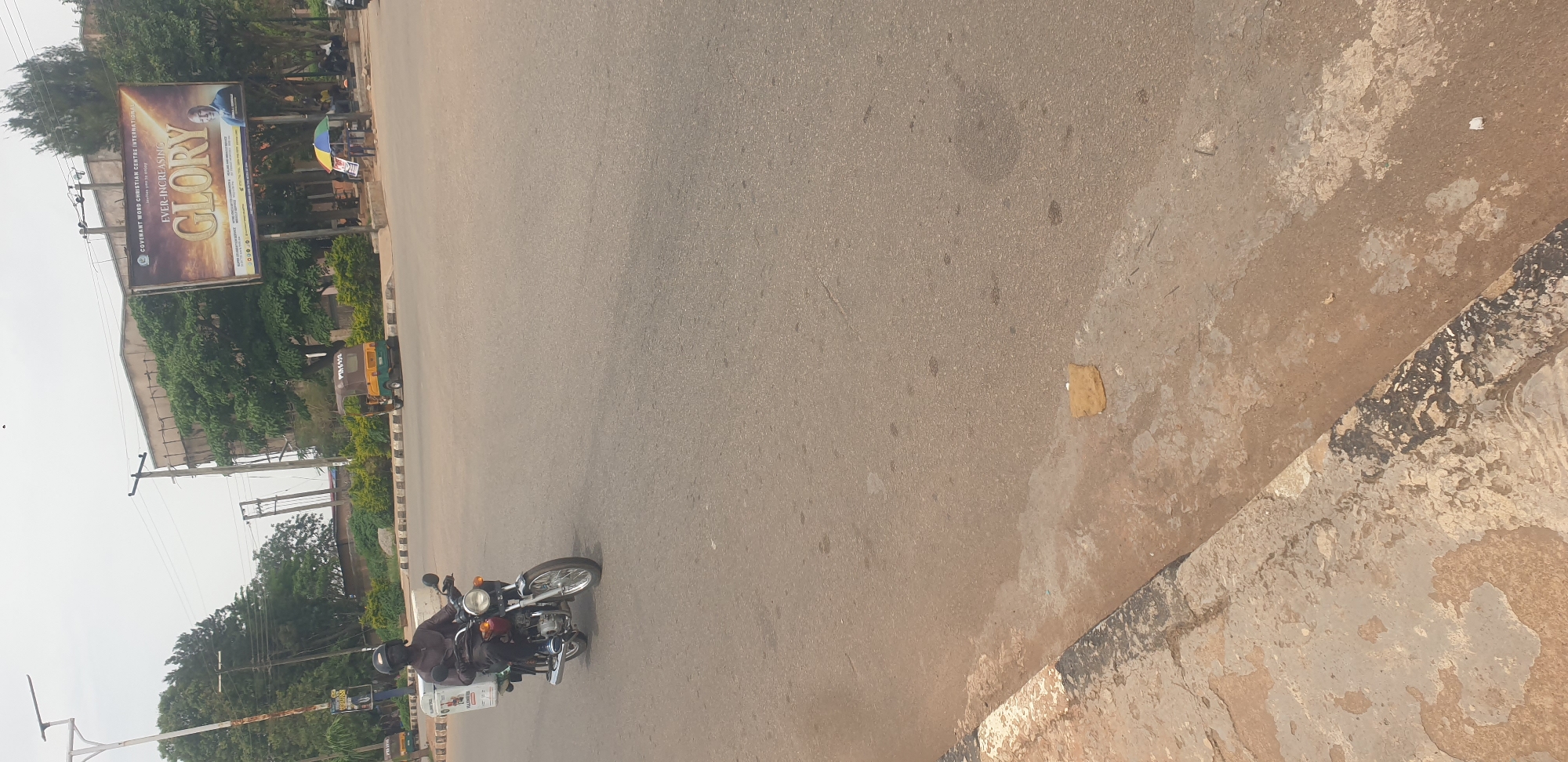 In Barakin Lerawi, Nigeria, a man wearing a dark helmet and a brown long-sleeved shirt rides a motorcycle on a paved road. The motorcycle has a white cargo box mounted on the rear. To the left of the motorcycle, there is a curb with black and white markings. In the background, a large billboard displays the text "COVENANT WORD CHRISTIAN CENTRE INTERNATIONAL" and "EVER-INCREASING GLORY" with an image of a man. Several trees are visible behind the billboard, along with some utility poles and wires. A yellow and green auto-rickshaw is parked on the side of the road, and some people are gathered under a colorful umbrella. The sky is overcast.