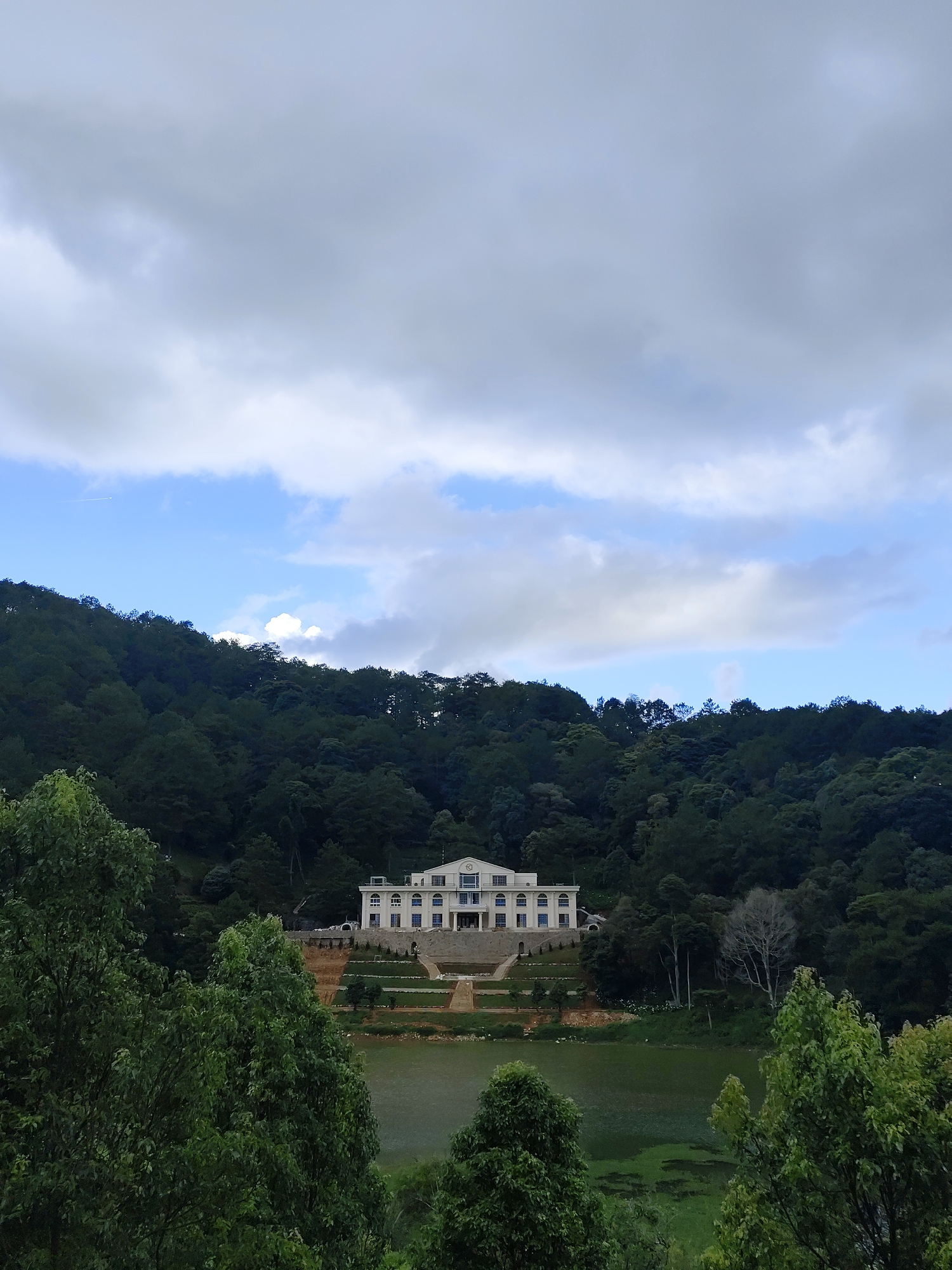 The image depicts a grand, light-colored villa situated on a terraced hillside overlooking a lake. The villa has a symmetrical facade with multiple windows and a central entrance. Terraced gardens lead from the base of the villa down to the lake's edge. The lake appears slightly murky with some algal growth visible. The hillside surrounding the villa is densely forested with lush, green trees. In the foreground, trees partially obscure the view of the lake and villa, creating a natural frame. The sky above is partly cloudy, with patches of blue sky visible between the clouds. The scene suggests a serene and possibly secluded location. This appears to be the Dinh II palace located in Da Lat, Vietnam.