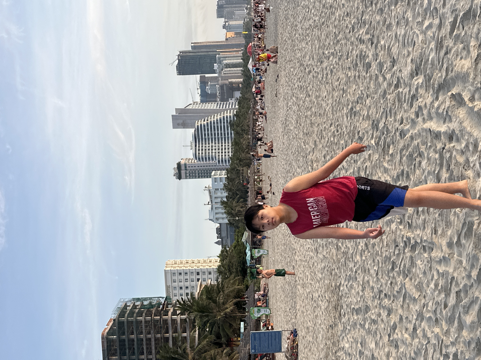 The image depicts a crowded beach scene in Da Nang, Vietnam, with the city's skyline in the background. In the foreground, a young person in a red tank top and patterned shorts walks towards the camera on the sandy beach. The beach is populated with numerous people, some relaxing on the sand, others walking, and some gathered under umbrellas. Buildings, including hotels and high-rise structures, some of which appear to be under construction, form the backdrop against a partly cloudy sky. Palm trees are scattered along the treeline behind the beach. The overall atmosphere suggests a bustling tourist destination.