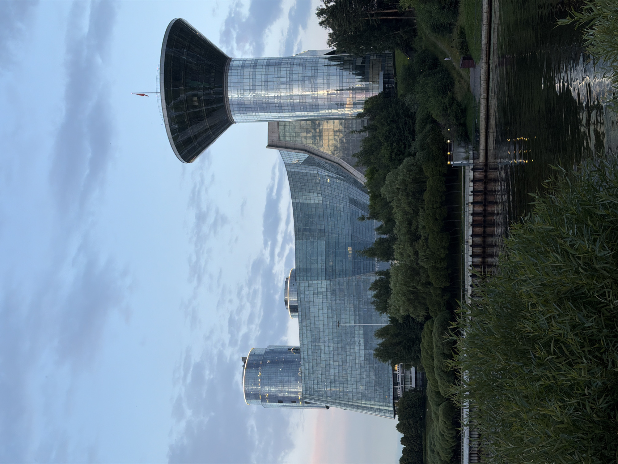 In Khimki, Russia, a striking architectural complex dominates the scene, set against a soft, cloudy sky. The primary structure is a multifaceted glass building with a distinctive curved facade, reflecting the light of the sky and surrounding environment. To the right, a cylindrical tower rises, topped with a large, inverted cone-shaped structure, from which a small red flag flutters. The building's glass exterior is illuminated with subtle, warm lights along its upper edges, adding to its modern aesthetic.
In front of the complex, a dense line of lush green trees forms a natural barrier, separating the building from a body of water. The water's surface is calm, reflecting the lights from the buildings and the sky, with ripples disturbing the otherwise smooth expanse. Along the water's edge, a reinforced concrete embankment with vertical pilings can be seen, suggesting a functional or historical element to the waterfront. Further back, a sloped grassy area is visible, dotted with more trees and a path leading towards a small structure. The overall impression is one of contemporary architecture harmonizing with a natural, waterside environment. There are no people clearly visible in the frame, but the presence of lighting and the structure itself implies human activity.