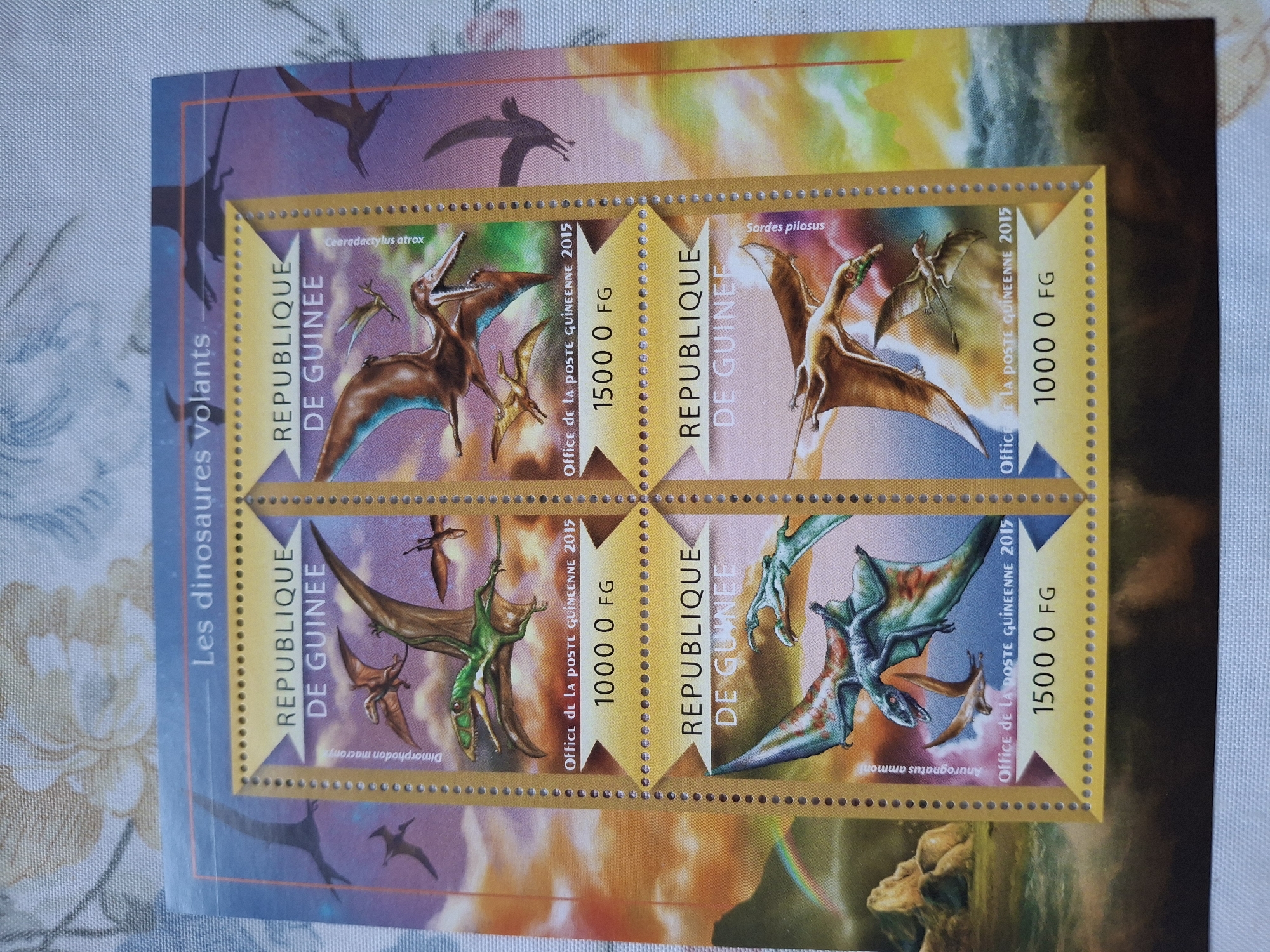 The image shows a set of four Guinea postage stamps featuring flying dinosaurs. The stamps are arranged in a 2x2 grid. Each stamp depicts a different species of flying dinosaur: Cearadactylus atrox (top left), Sordes pilosus (top right), Dimorphodon macronyx (bottom left), and Anurognatus ammoni (bottom right). The stamps are labeled "REPUBLIQUE DE GUINEE" and feature the denomination, "Office de la POSTE GUINEENNE 2015." The surrounding background has an illustration of multiple flying dinosaurs. The image appears to be taken on a patterned surface. There is no information to suggest this image has anything to do with the city of Pleven, Bulgaria.