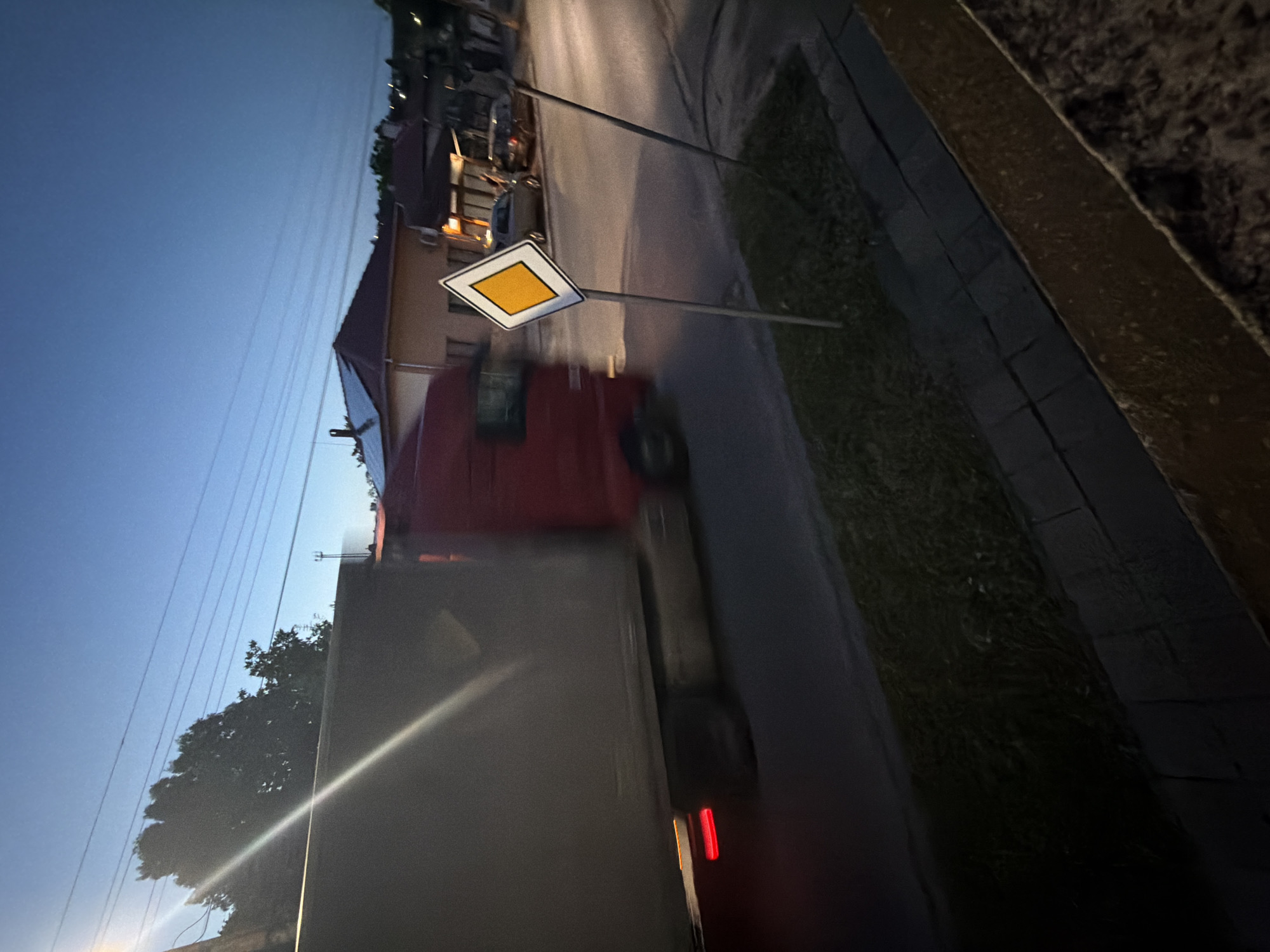 The image depicts an evening scene on a road in Iskar, Bulgaria. A large, grey/silver truck with a red cab is prominently featured, driving along the asphalt road. Motion blur indicates the truck is in movement, and its red tail lights are visible.
To the right of the truck, a distinct yellow square road sign with a white border is posted on a pole, signifying a "priority road" or "main road." The road itself is dark asphalt, bordered by a grassy verge on the left. In the background, several houses with visible roofs are scattered, suggesting a residential or semi-rural setting.
The sky is a deep blue, characteristic of dusk or early night, with multiple power lines or utility wires stretching across it. Trees are also visible in the distance. The right side of the frame is bordered by a dark, textured wall or structure. No people are visible, and there is no discernible text other than the universally recognized road sign.