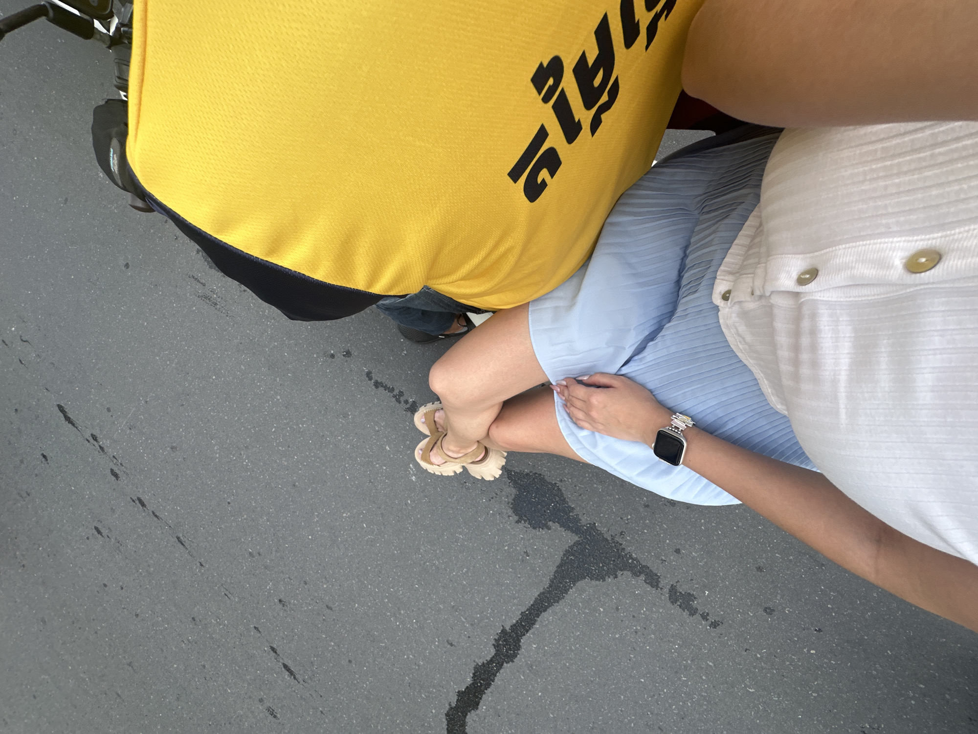 The image shows a person sitting on a motorcycle in what appears to be an urban environment. The person is wearing a white ribbed top and a light blue pleated skirt. They have a smartwatch on their left wrist. To the right of the person is a large, bright yellow bag with black text in what appears to be Khmer script. The motorcycle is on an asphalt road with some dark wet patches. The background is blurred, but suggests movement. The image quality is good, with clear details of the subjects and their clothing.