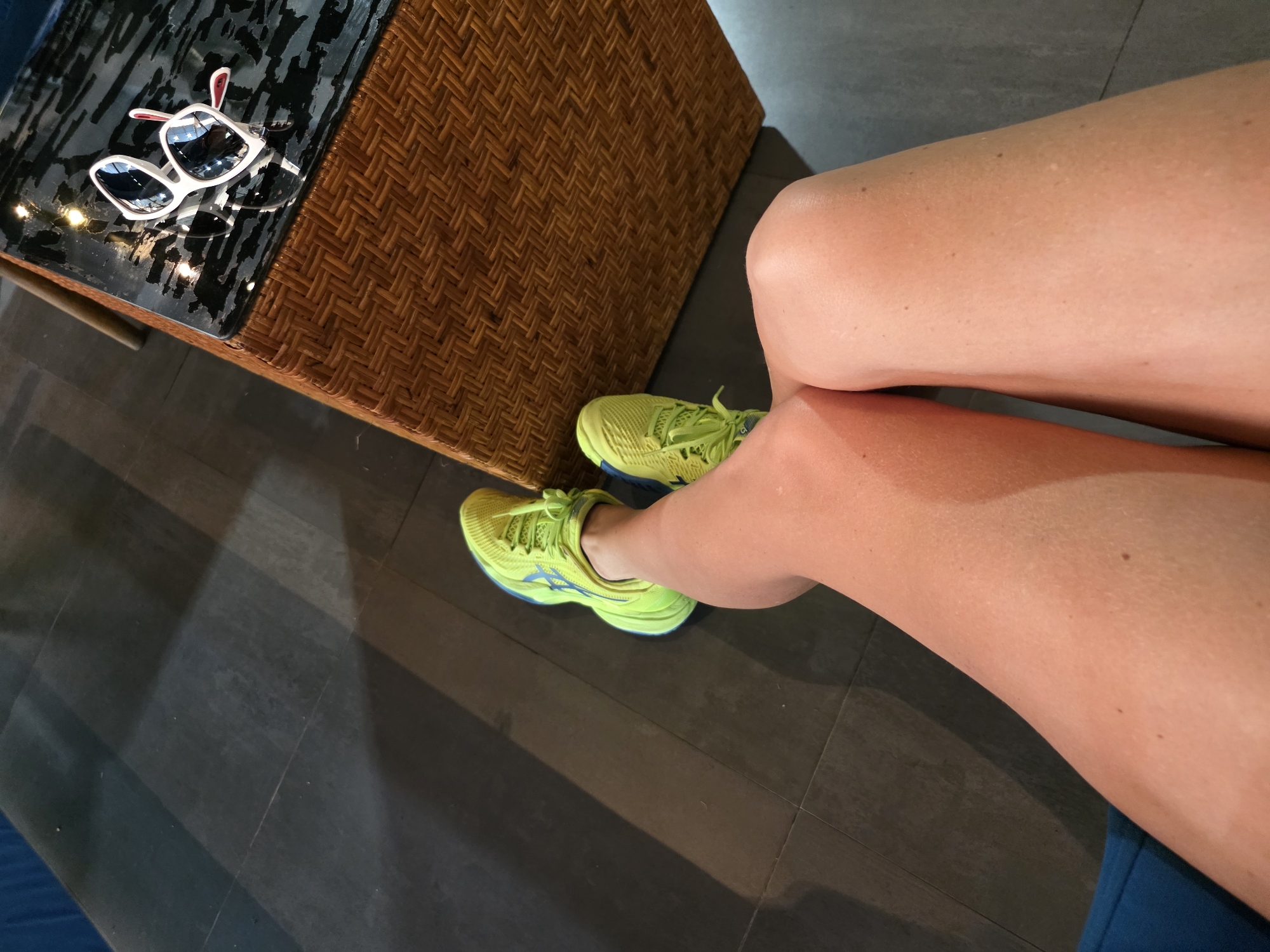 A close-up, high-angle shot shows a person's legs and feet, as well as a wicker coffee table with sunglasses on it, in what appears to be an indoor seating area. The floor is made of large, dark grey tiles with a subtle pattern. The person is wearing bright lime green athletic shoes with blue accents. The legs appear tan and smooth. The coffee table is rectangular with a woven wicker base and a glossy black top, on which a pair of white-rimmed sunglasses with dark lenses and red accents on the temples are resting. The sunglasses reflect the surrounding environment. The overall lighting suggests an indoor setting with artificial light sources. This scene could be in a hotel lobby, resort lounge, or a similar establishment in the tourist area of North Kuta, Indonesia, given the tropical-inspired furniture and the presence of sunglasses, common in such locations.