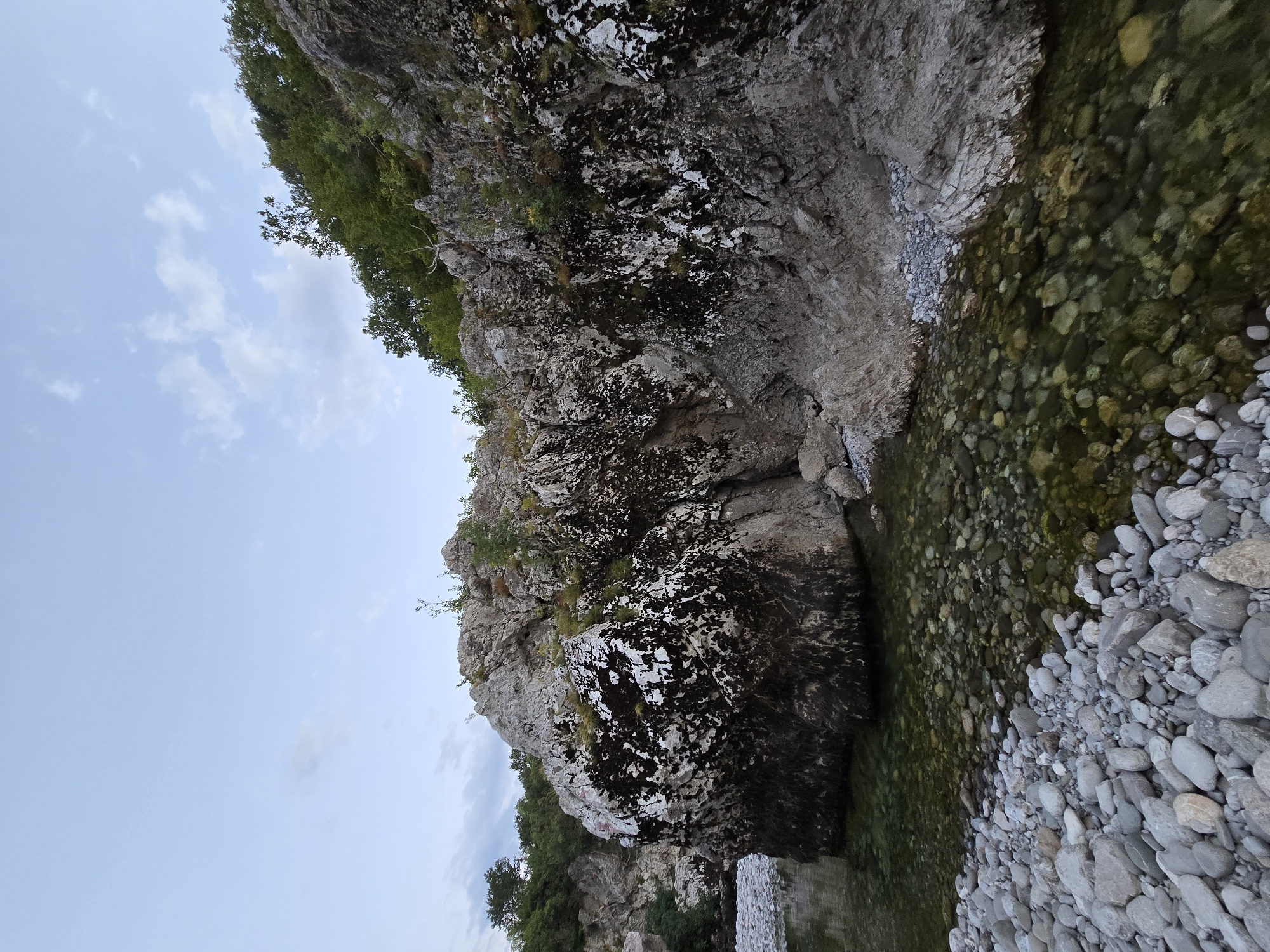 The image depicts a rocky riverbed with clear, shallow water flowing over smooth, grey stones. A large, weathered boulder dominates the left side of the frame, its surface mottled with dark and white lichens. The river curves to the left, disappearing behind the boulder. The right side of the scene is a steep, rocky embankment covered in vegetation, with trees and shrubs growing from crevices. The sky above is a pale blue with scattered white clouds, suggesting either early morning or late afternoon. The overall impression is of a natural, untouched landscape. The location is identified as Bioče, Montenegro, a settlement situated in a mountainous region known for its rivers and natural beauty.