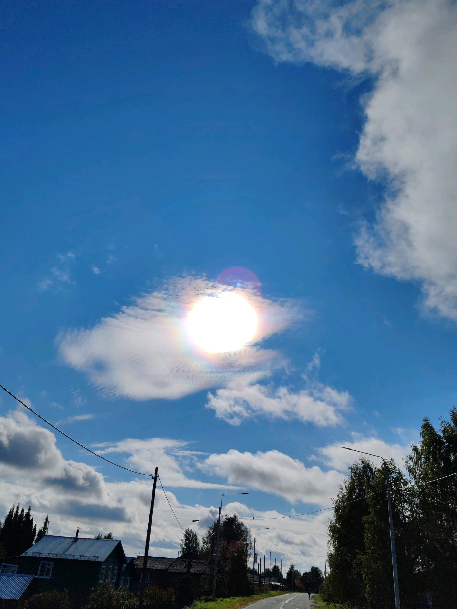 This image depicts a rural scene in Chukhlem, Russia, under a bright blue sky dotted with fluffy white clouds. A prominent solar halo, a rainbow-like ring around the sun, is visible in the upper part of the frame, suggesting the presence of ice crystals in the atmosphere. Below the sky, a winding asphalt road stretches into the distance, with a lone figure walking on its shoulder. To the left of the road, several houses with dark roofs and visible windows are nestled amongst trees. A utility pole with wires crisscrosses the sky, and streetlights line the road, indicating a populated area. The overall impression is one of a serene, possibly autumnal, day in a Russian village.