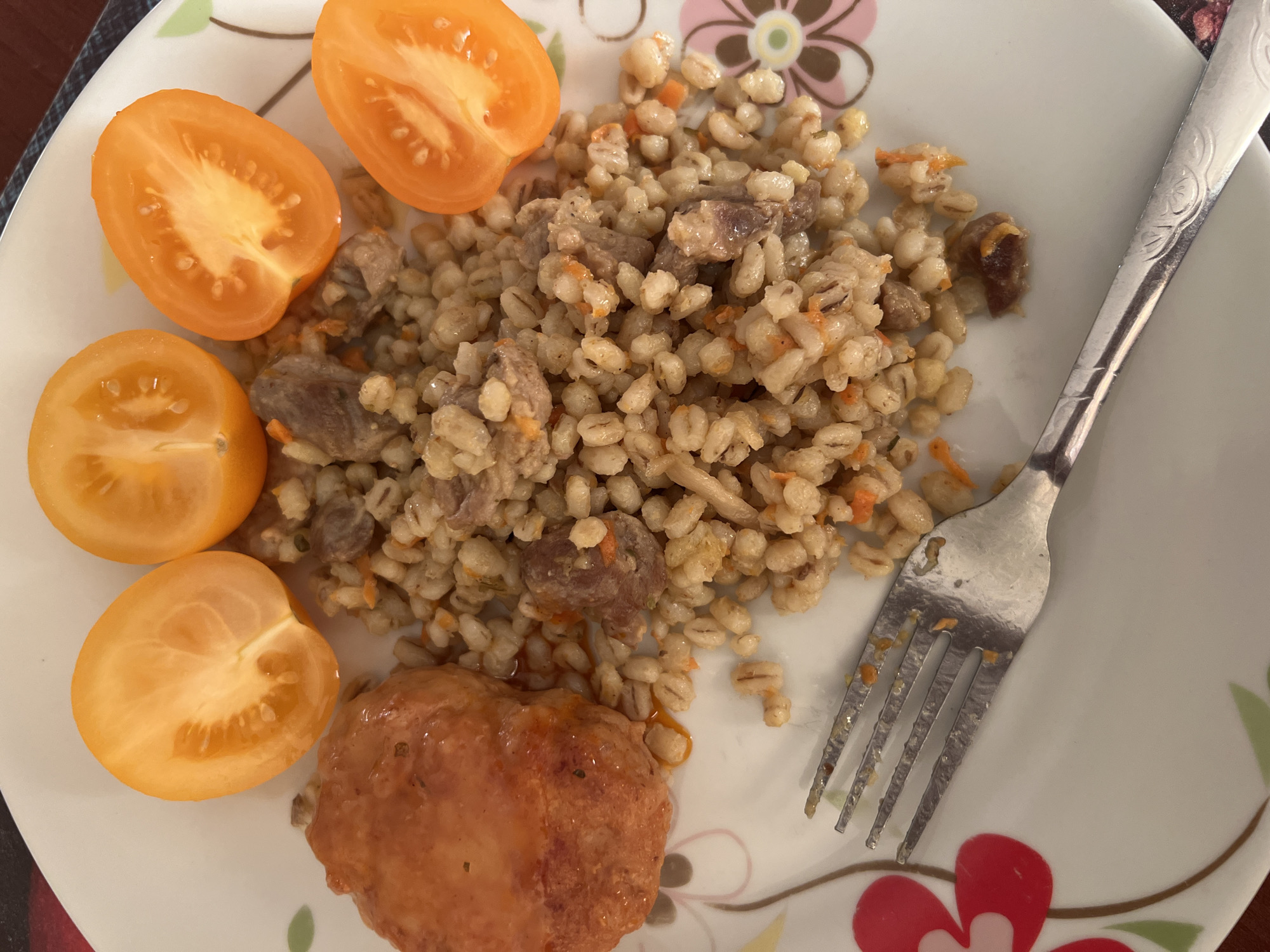 A top-down, close-up shot shows a plate of food, likely served in a home dining setting in Minsk, Belarus. The plate is white with a floral design around the rim, featuring pink and red petals. A silver fork rests in the lower left quadrant of the plate.
The food consists of a portion of barley or similar grain, mixed with small pieces of meat and visible shreds of orange carrot. To the left of the grain mixture, there is a golden-brown, round item, possibly a croquette or a piece of fried chicken, coated in sauce. At the top of the plate, four wedges of orange tomato are arranged, each cut in half. The overall presentation suggests a home-cooked meal. The lighting is consistent with indoor ambient light.