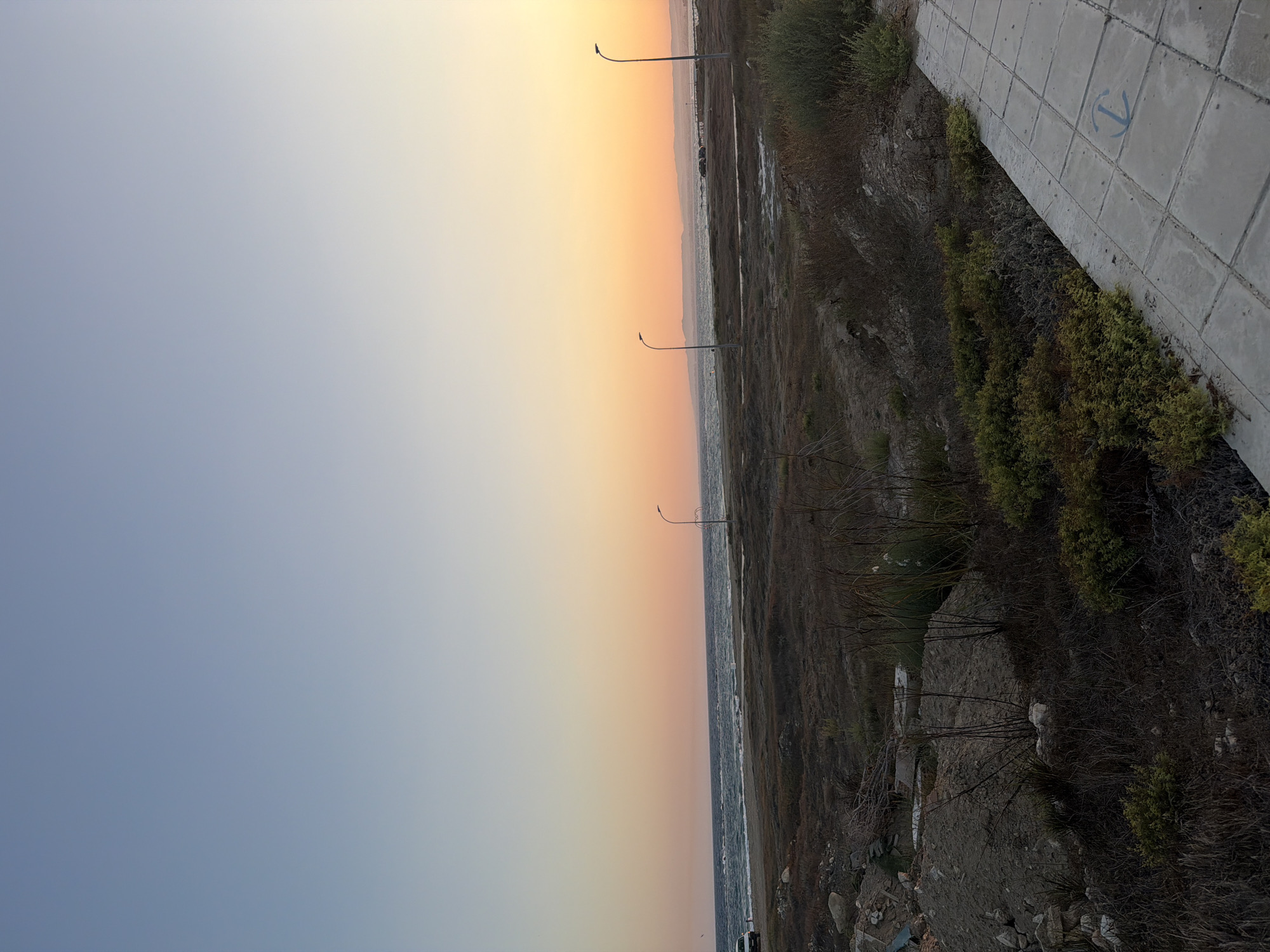 The image depicts a coastal scene at sunset in Meneou, Cyprus. The sky displays a gradient of soft colors, transitioning from a pale blue at the bottom to a warm orange and yellow near the horizon, indicating the sun is setting. The sea is visible as a dark, wavy expanse.
On the right side of the image, a paved walkway or road runs diagonally, constructed of rectangular concrete tiles. A blue anchor symbol is visible on one of these tiles. Adjacent to the walkway, the landscape is covered in dry, low-lying vegetation and shrubs. Two streetlights with curved poles stand along the edge of the land, one closer to the viewer and another further down the coast.
In the distance, along the horizon, a thin line of land or buildings can be seen. The overall impression is one of a quiet, natural setting, perhaps a seaside promenade or a coastal path. The time of day is clearly sunset, and the weather appears calm. No people are visible in the frame.