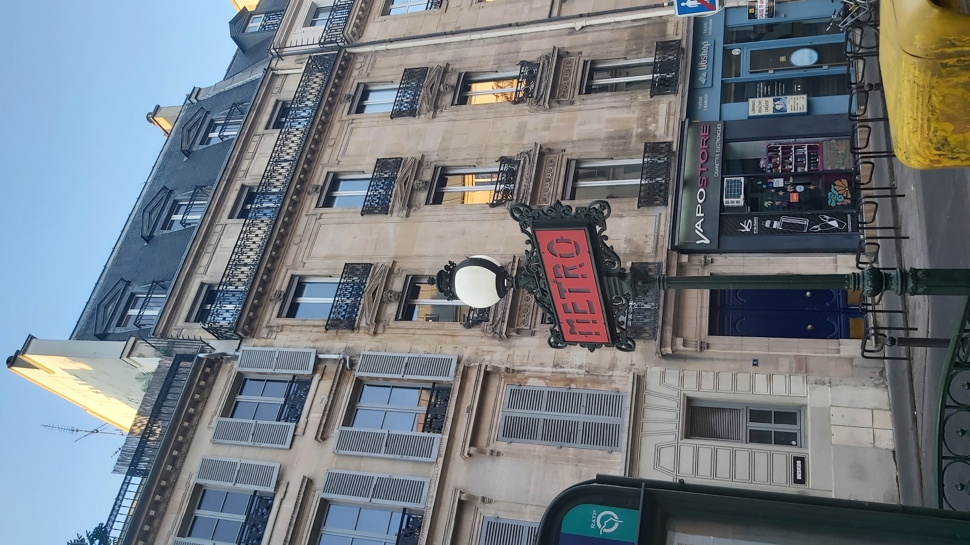 The image shows a street scene in Paris, France, with a Metro station sign prominently displayed. The sign, a classic red "METRO" lettering within an ornate green metal frame, is mounted on a lamppost topped with a globe light. Behind the sign, a multi-story Haussmann-style building dominates the background, characterized by its beige stone facade, numerous windows with grey shutters, and decorative wrought-iron balconies. The upper floors feature mansard roofs with dormer windows. Below the building's upper floors, a row of shops is visible at street level. One shop has a dark facade with the text "VAPO STORE" and "CIGARETTES ELECTRONIQUES" displayed, suggesting it sells vaping products. Next to it, a shop with a blue facade shows signage indicating "LIBANAISE HEALTHY CREATIF," implying it offers Lebanese cuisine or related services. The street in front of the shops is paved, and several black bicycle racks are positioned along the sidewalk. A portion of a yellow object, possibly a vehicle, is visible in the lower right corner of the image. The sky above is clear and a pale blue, indicating daylight, likely late afternoon or early morning due to the warm lighting illuminating the building. The overall impression is of a typical Parisian streetscape with a functional, well-maintained urban environment.