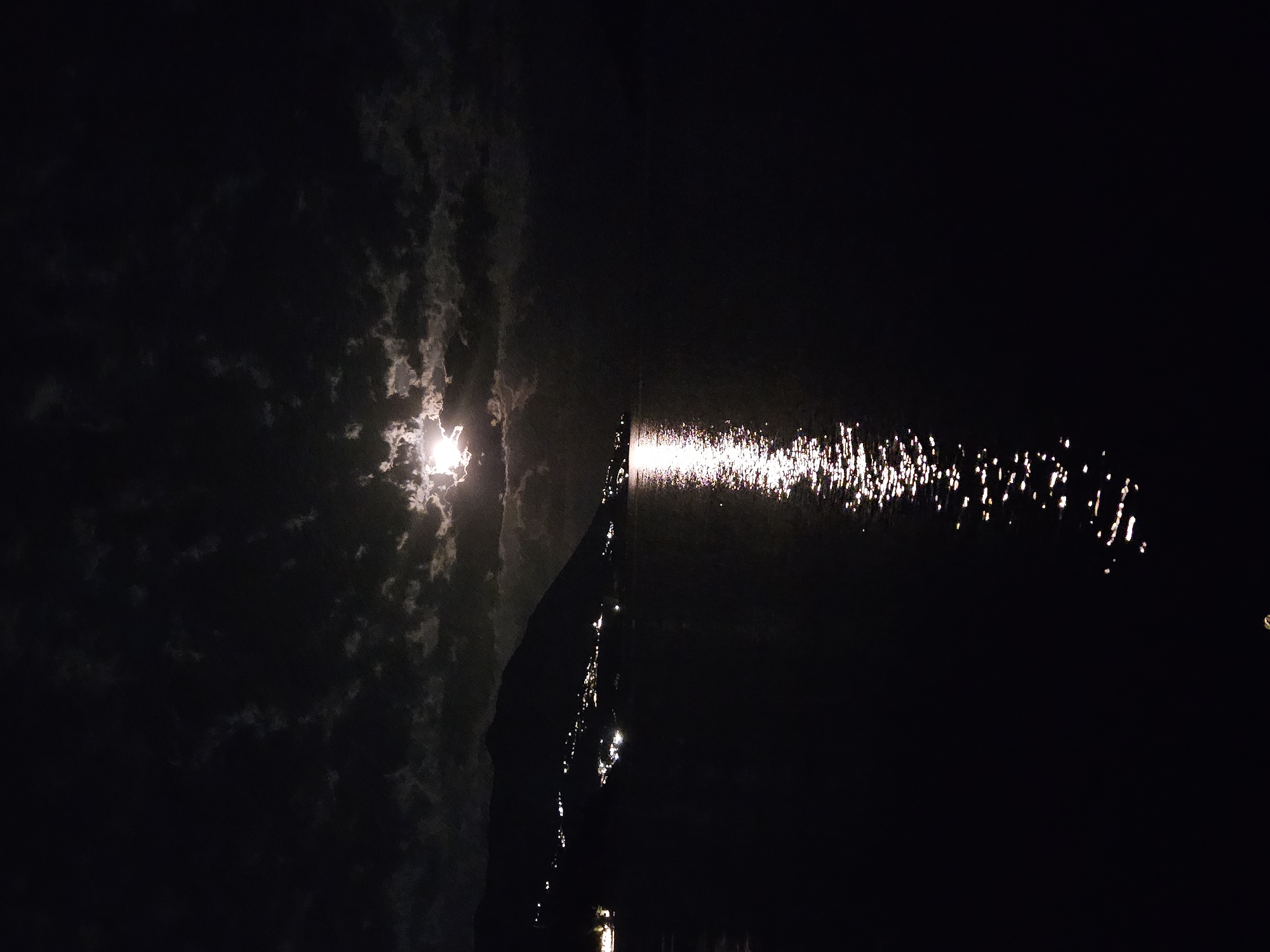 The image displays a high-contrast nocturnal scene, dominated by the moon and its reflection on a body of water. On the left side, a bright, intense moon shines through broken, dark clouds, casting a soft glow on their edges.
Extending horizontally from the approximate position of the moon, across the center of the frame and towards the right, is a shimmering path of moonlight reflecting on the water's surface. This reflection is not a solid beam but appears as numerous sparkling, elongated specks and streaks of light, indicating a slightly rippled or moving water.
A distinct, dark, vertical silhouette stands in the mid-ground, positioned between the moon and the more extensive part of its reflection. This object is thin and tall, resembling a boat's mast or a slender structure. The sky above the moon and the water surface beyond the main reflection are enveloped in deep darkness.
The time of day is clearly night. No specific weather conditions other than partial cloud cover are apparent. No people, discernible man-made objects beyond the potential mast, or visible text are present. The scene conveys a sense of peaceful solitude and natural beauty under the moonlight. The location appears to be an open body of water.