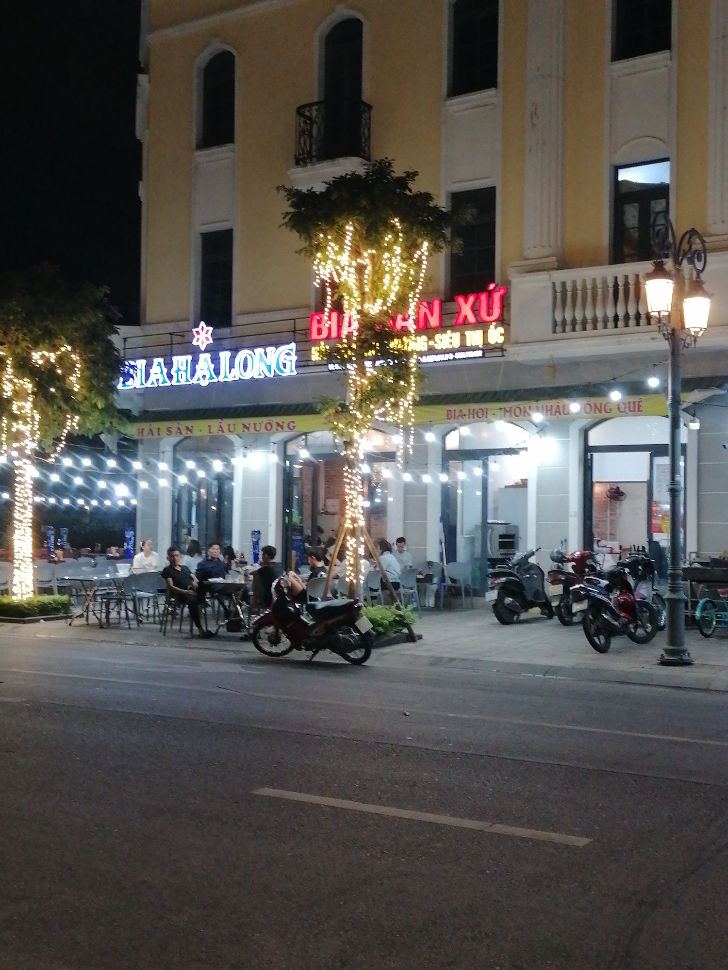 The image captures a vibrant nighttime scene at an outdoor restaurant or bar in Bai Chay Ward, Vietnam. A multi-story building with a light yellow facade and distinctive arched windows forms the backdrop. Overhead, a large illuminated sign displays "BIA HẠ LONG" (Bia Ha Long) with a red star symbol, and partially visible text "ẨN XỨ SIÊU THỊ ỐC". A yellow banner below further advertises "HẢI SẢN - LẨU NƯỚNG" (Seafood - Hotpot/Grilled Food) and "BIA HƠI - MÓN NHẬU ÔNG QUÊ" (Draft Beer - Countryside Snacks/Dishes).
The outdoor dining area is active with several people seated at tables, socializing under an array of string lights. These lights also adorn the trunks of nearby trees, creating a festive and inviting atmosphere. Numerous motorbikes are parked on the sidewalk, reflecting typical urban transportation in Vietnam. A classic street lamp with two lanterns stands on the right, contributing to the ample illumination of the scene. The street in the foreground is dark, indicating it is late evening or night. The overall impression is one of a lively and casual social gathering.