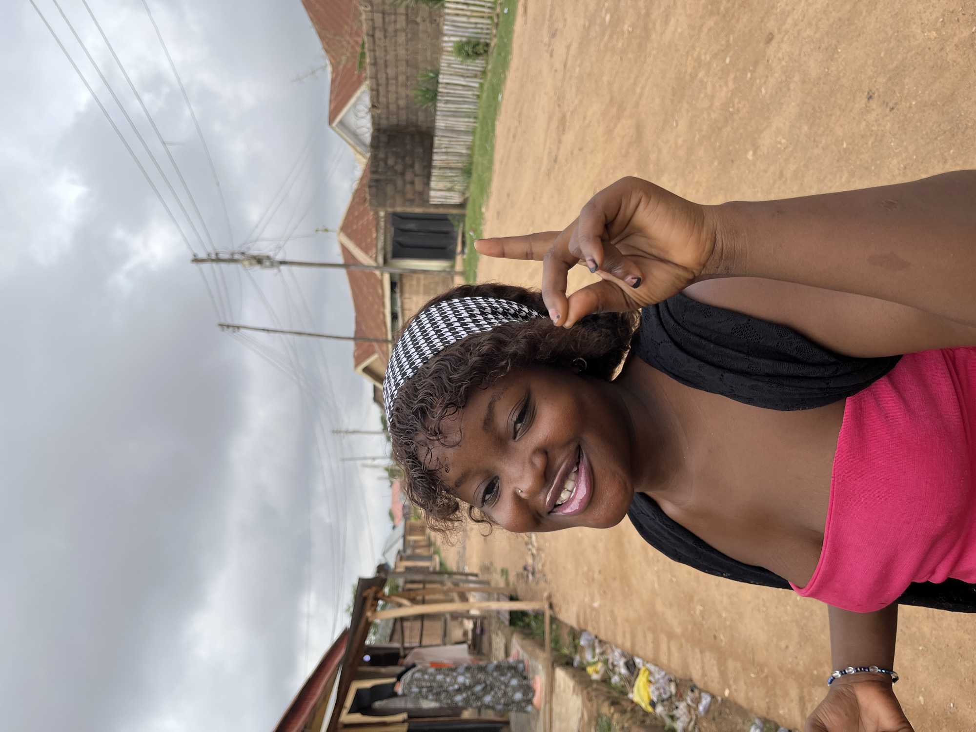 A cheerful young woman is captured in a close-up outdoor portrait, smiling broadly and making a peace sign with her right hand. She has dark, curly hair adorned with a black and white houndstooth headband, a nose piercing, and dark nail polish. She is wearing a vibrant hot pink sleeveless top under a black garment and a beaded bracelet on her left wrist.
The setting is an unpaved, dusty road in what appears to be a residential or semi-urban area in Barakin Akawo, Nigeria. In the background, simple buildings with brick walls and corrugated metal roofs are visible, along with wooden fences. The sky is overcast and grey, with utility poles and numerous power lines stretching across the upper portion of the frame. Some scattered litter, including plastic bags, can be seen on the roadside. The image depicts a bright, cloudy day. There is no visible text.