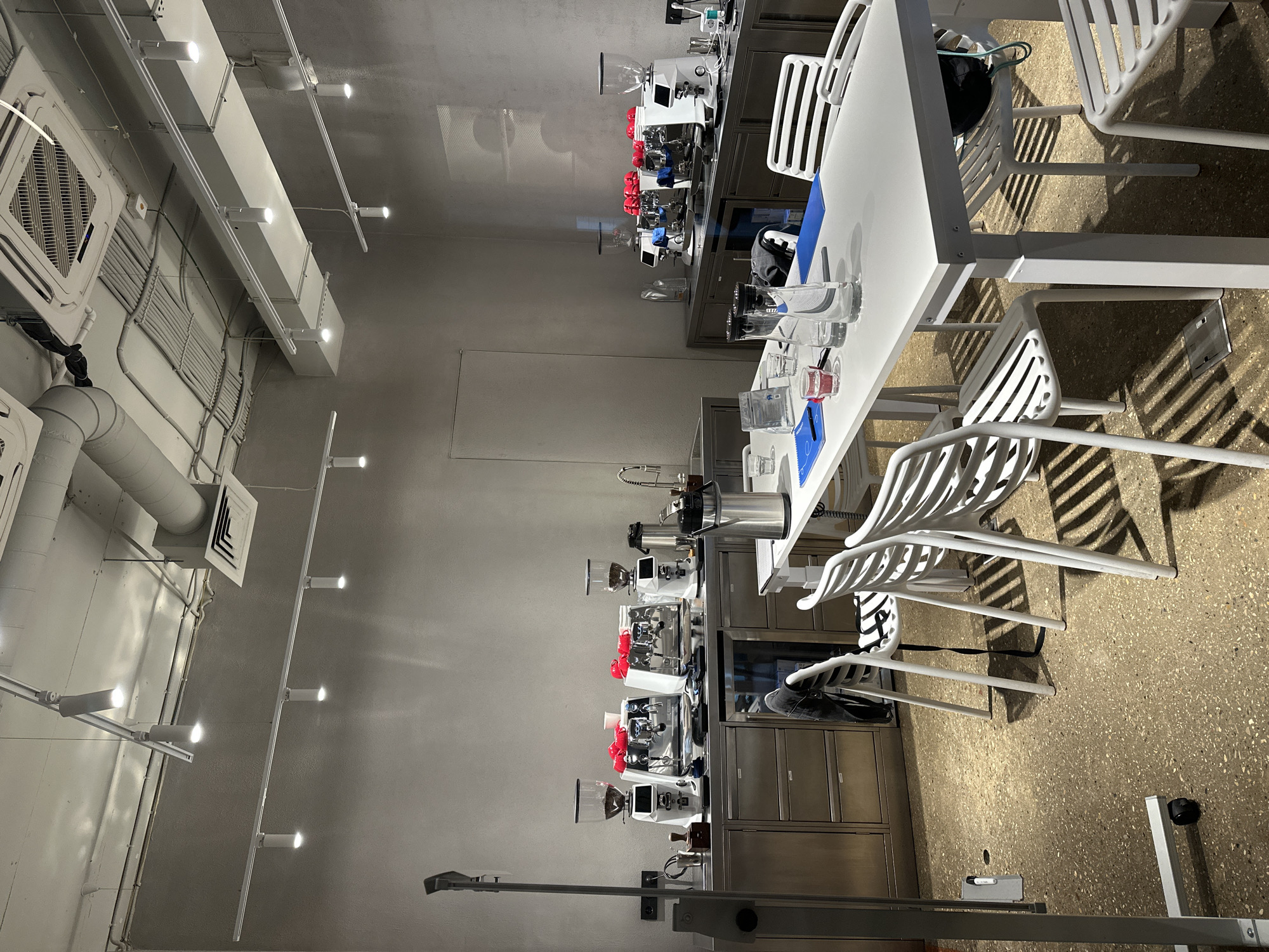 The image shows a modern, clean, and well-lit interior space, likely a cafe or a barista training facility. Several rows of professional-grade espresso machines and grinders are neatly arranged on stainless steel counters, suggesting a setup for coffee preparation or education. White tables and chairs, also in a contemporary design, are placed around the space, some with shadows cast by overhead lighting.
On one of the tables, there are glasses, a carafe of water, and what appears to be a blue placemat or cutting board. The overall aesthetic is minimalist and functional, with a focus on hygiene and organization. The lighting is bright and consistent, indicating it is daytime or that the artificial lighting is sufficient to illuminate the space uniformly.
The walls are a neutral gray, and the floor is a speckled terrazzo material. Ductwork and electrical conduits are visible on the ceiling, contributing to the industrial yet refined look of the setting. There are no people visible in the image, giving the impression of an unoccupied space, perhaps between service hours or during a class preparation.
Notable details include the array of gleaming espresso machines, each with a glass hopper filled with coffee beans on top of the grinders. The chairs have a slatted design, adding to the visual texture of the scene. The image is shot from a high angle, providing a broad view of the setup. There is no visible text in the image.
