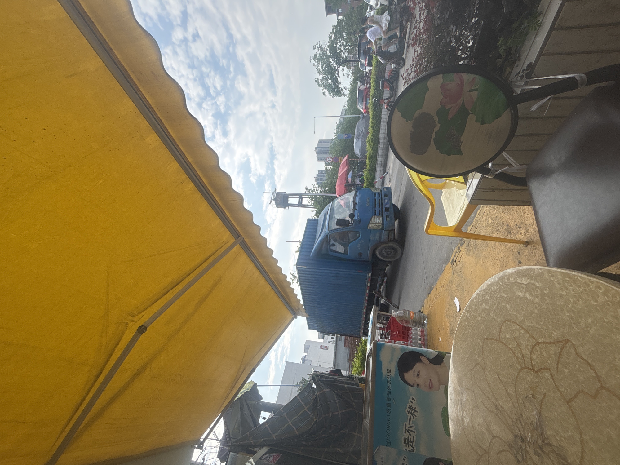 The image is a street-level view in Nanhai District, China, dominated by a large yellow awning casting shadows. In the background, a blue shipping container truck is driving on a street alongside other vehicles, including motorcycles and cars. Buildings and trees are visible in the distance. In the foreground, a round table with a floral pattern is partially visible, along with yellow and dark chairs. A poster or advertisement featuring a woman's face and Chinese text is also present. The scene appears to be a commercial or roadside area.