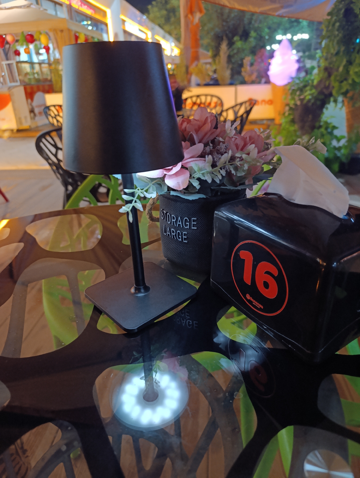 An outdoor table setting at night features a black lamp with a cylindrical shade, positioned behind a pot of artificial pink and white flowers. To the right of the flowers is a black tissue box dispenser with a red circle containing the number "16" and a small logo. The table has a reflective dark surface with an abstract pattern. In the background, out-of-focus elements include chairs, greenery, and illuminated structures suggesting a commercial area like a restaurant or cafe. The lighting is low, with the lamp and a bright circular reflection on the table being prominent light sources.