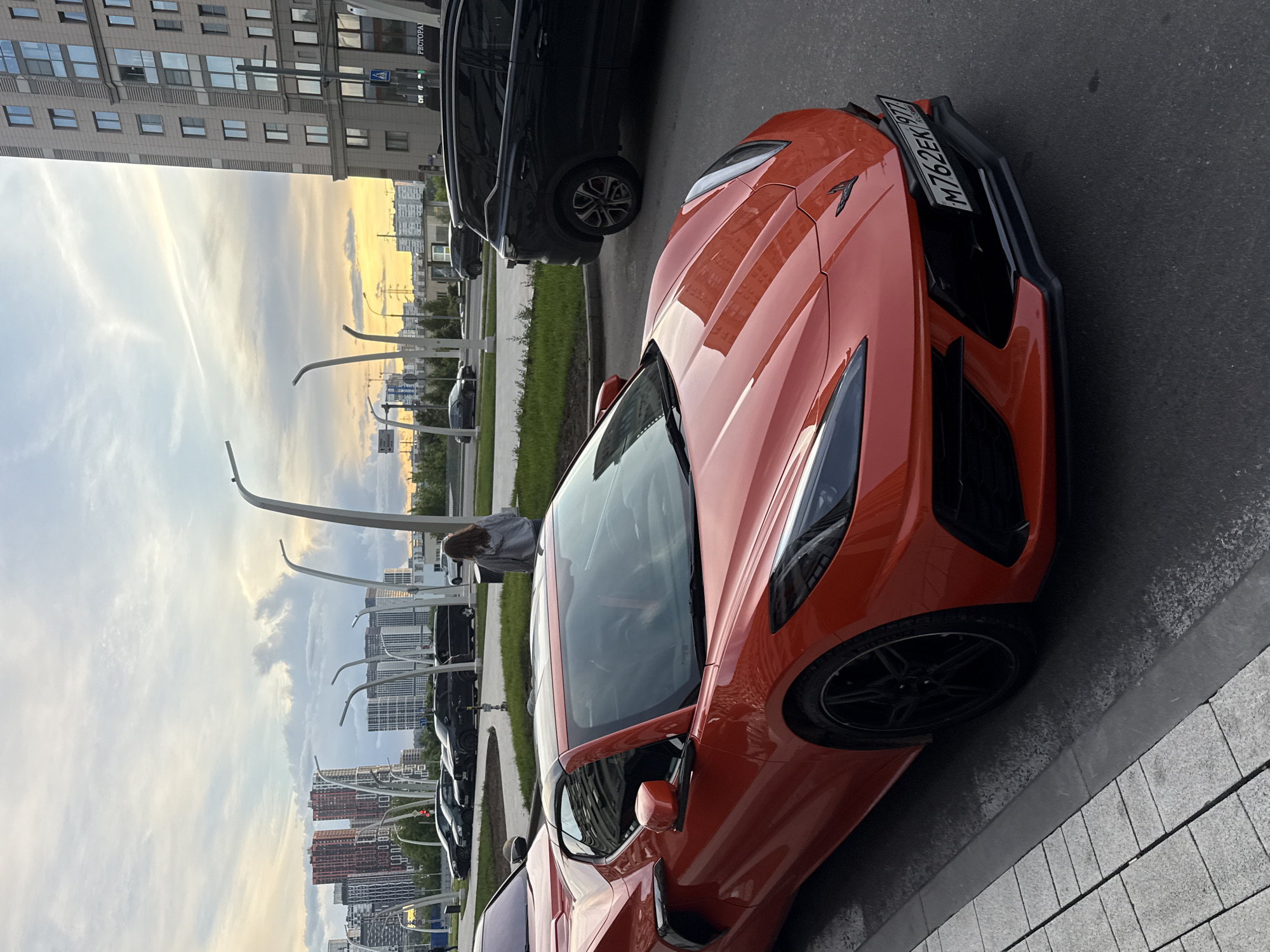 The image depicts an orange Corvette sports car parked on a city street in Moscow, Russia. The car is positioned on the right side of the frame, with its front end facing the viewer. To its left, there is a line of parked cars, including a black SUV. A woman is standing near the parked cars. The background reveals tall apartment buildings and a sky with a sunset glow. The license plate of the orange Corvette is visible, showing "M762EK 797". The setting appears to be an urban environment during the evening hours.