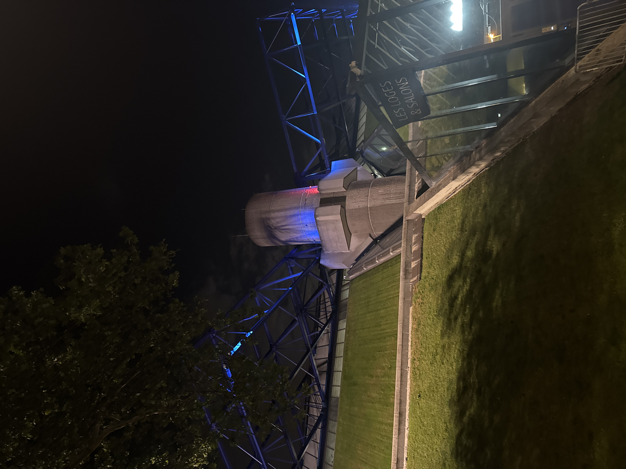 The image shows La Géode at night in Paris, France. It is a mirrored geodesic dome that is illuminated with blue and red lights. The structure is surrounded by a metal framework. To the right is a sign that reads "LES LOGES & SALONS." The image also includes trees, grass, and dark sky.