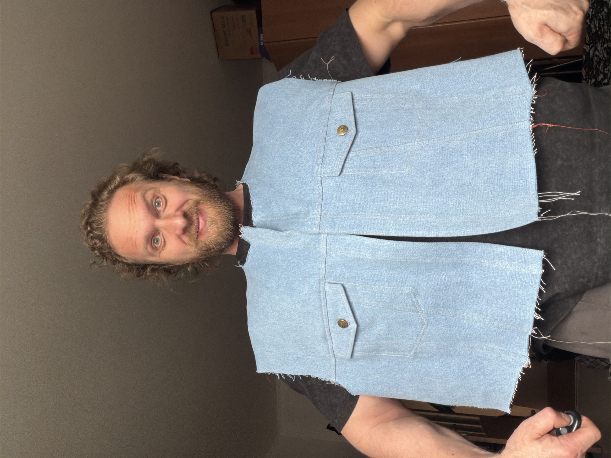 A man is holding a light blue denim vest up to his chest. The vest has two pockets with buttons. The fabric of the vest is frayed around the edges. The man has curly hair, a beard, and light eyes. He is wearing a dark grey t-shirt. The background includes a grey wall, a wooden cabinet, and a cardboard box.