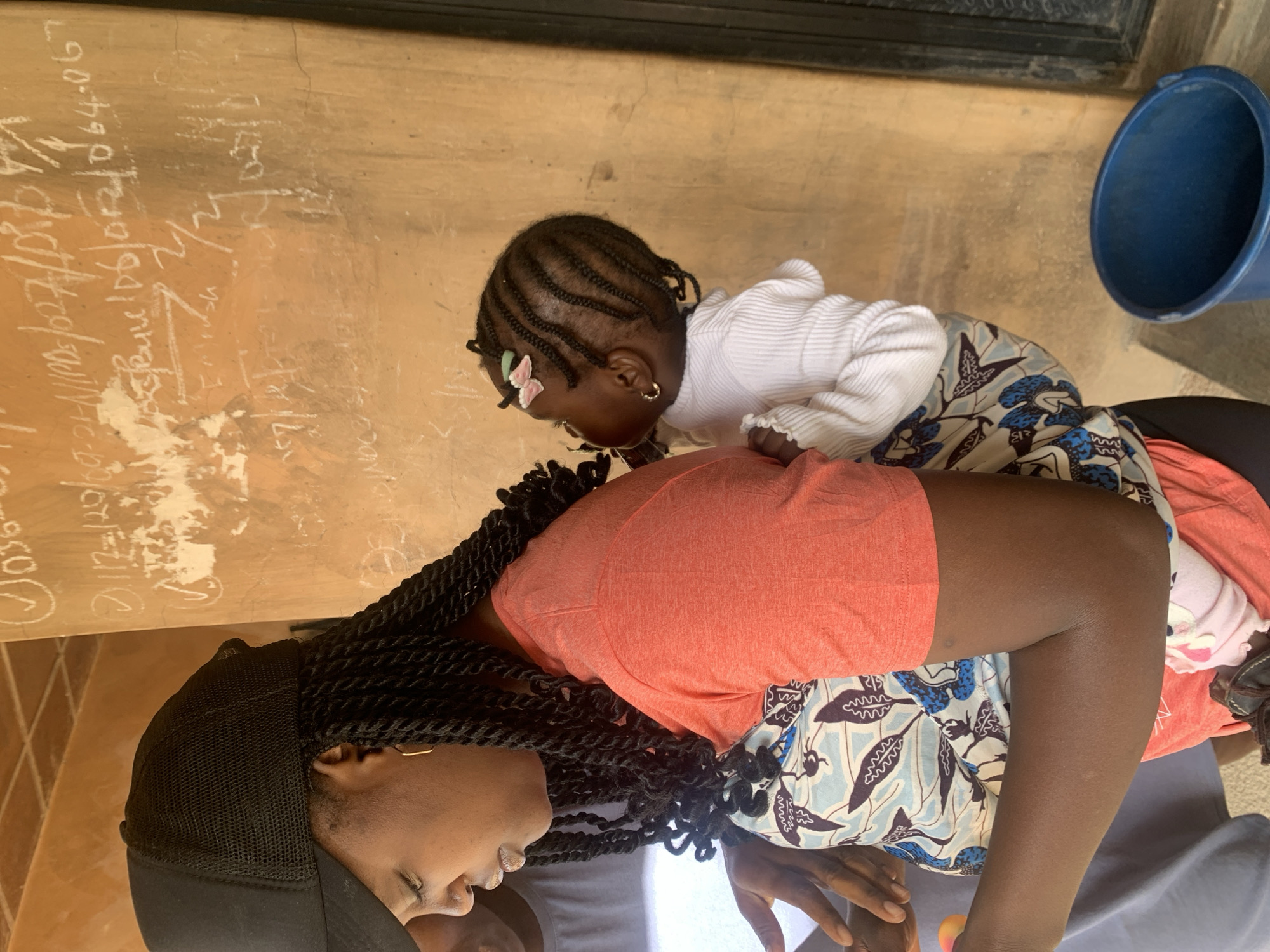 A side profile of a Black woman wearing a black trucker hat and an orange t-shirt is shown. Her dark hair is in numerous braids, some of which fall over her shoulder. She is carrying a young Black child on her back, who is wearing a white long-sleeved shirt and is facing away from the camera. The child has braided hair with a pink bow. They are positioned in front of a textured, light brown wall with writing and drawings on it, characteristic of an outdoor area in Jos, Nigeria. A blue plastic bucket is visible on the lower right side of the image. Another person, only partially visible, is to the left of the woman.