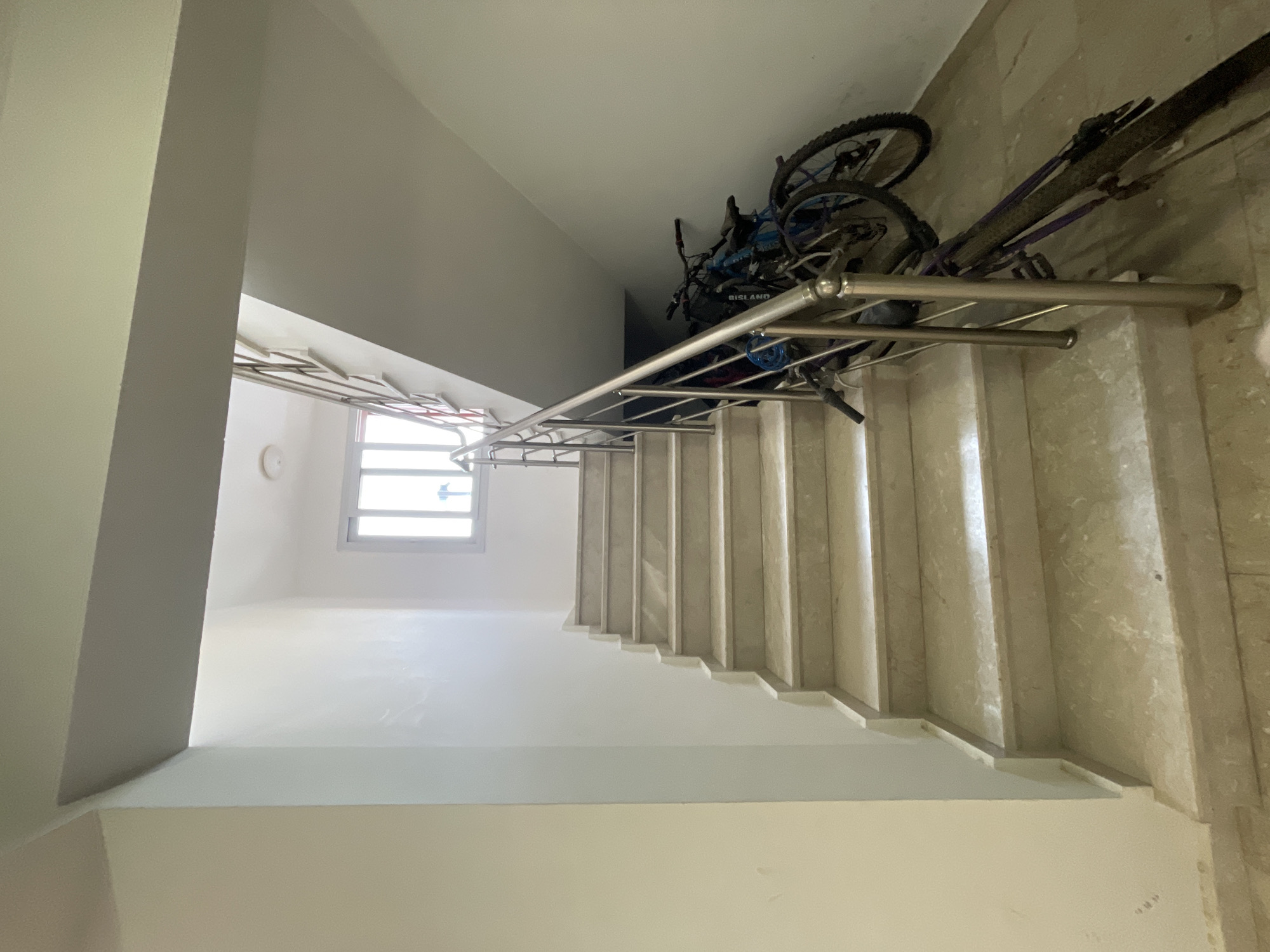 The image shows an indoor staircase with a marble finish. The staircase has a silver metal railing on the right side. At the bottom of the stairs, several bicycles are parked against the wall. There is a window with horizontal panes at the top of the staircase. The walls are painted white and a light beige. The lighting in the scene is artificial. The overall environment suggests a communal area within a residential building, possibly in Trikomo, Northern Cyprus.